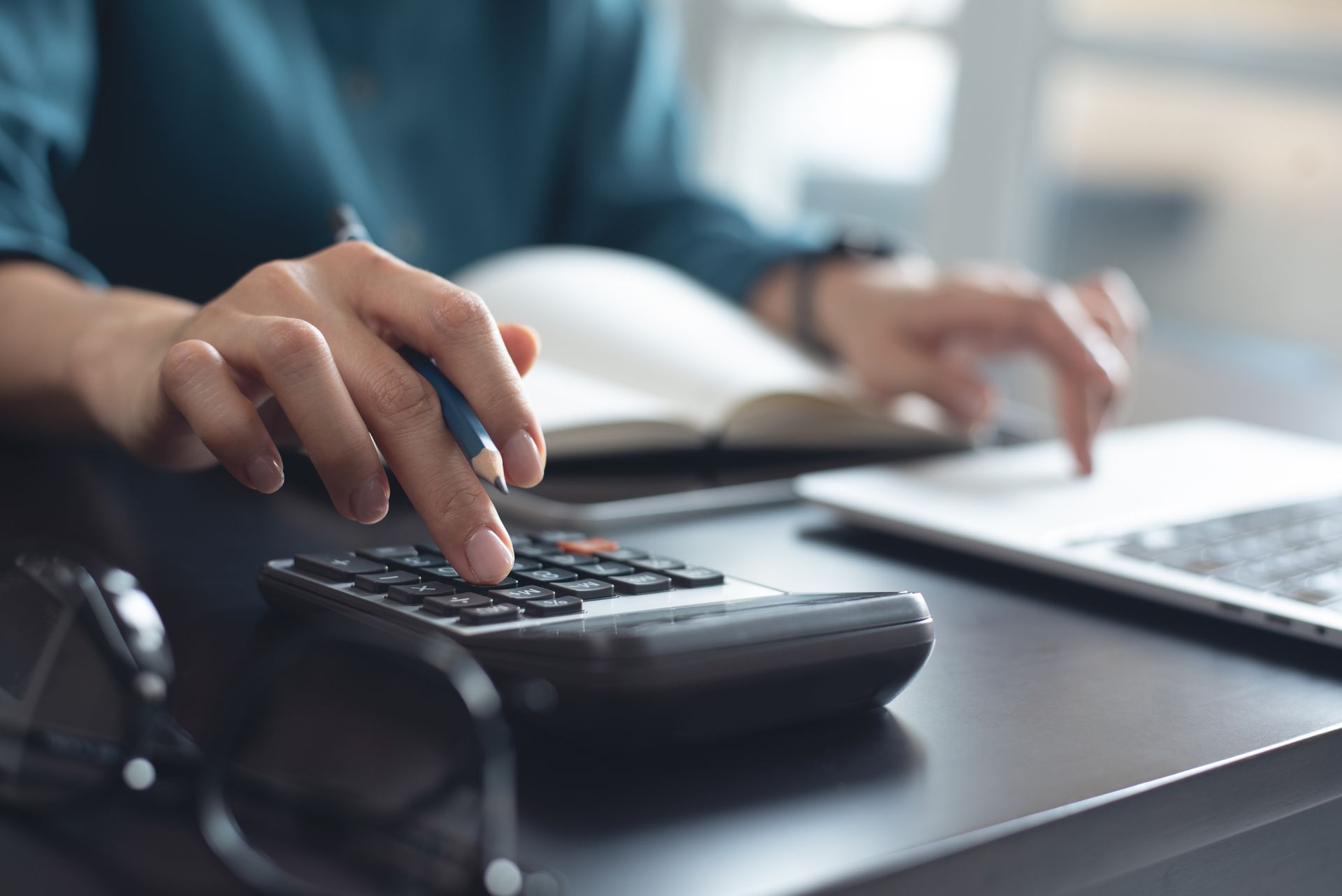 Person calculating on a calculator, with a laptop and open book on a desk. Person calculating on a calculator, with a laptop and open book on a desk.