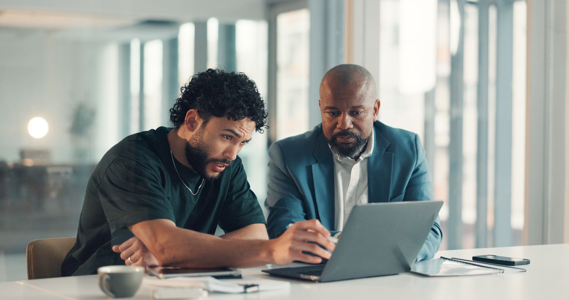 Two men looking at a laptop in a modern office, one pointing, discussing details.