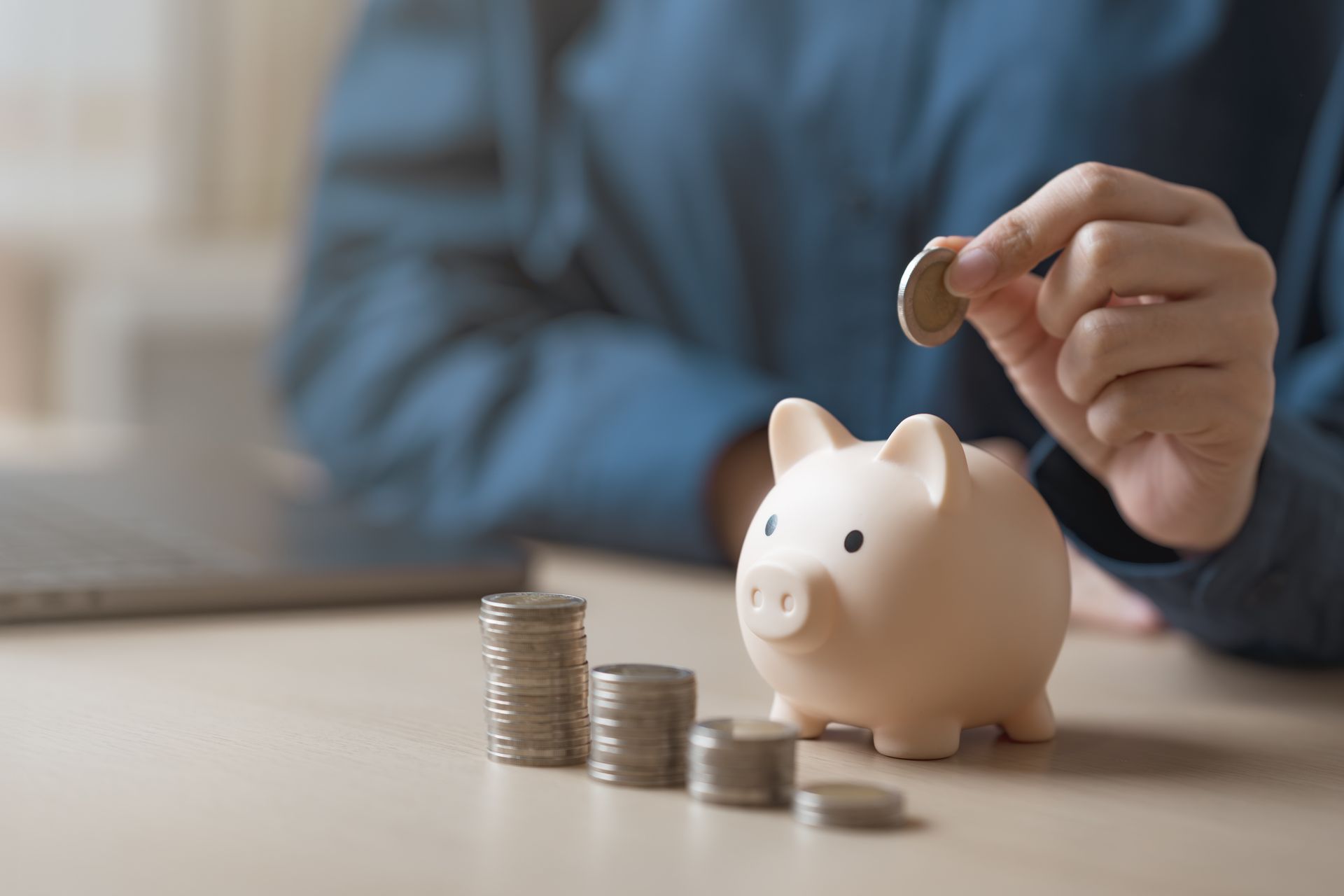 Person adding a coin to a piggy bank; stacks of coins on the table.