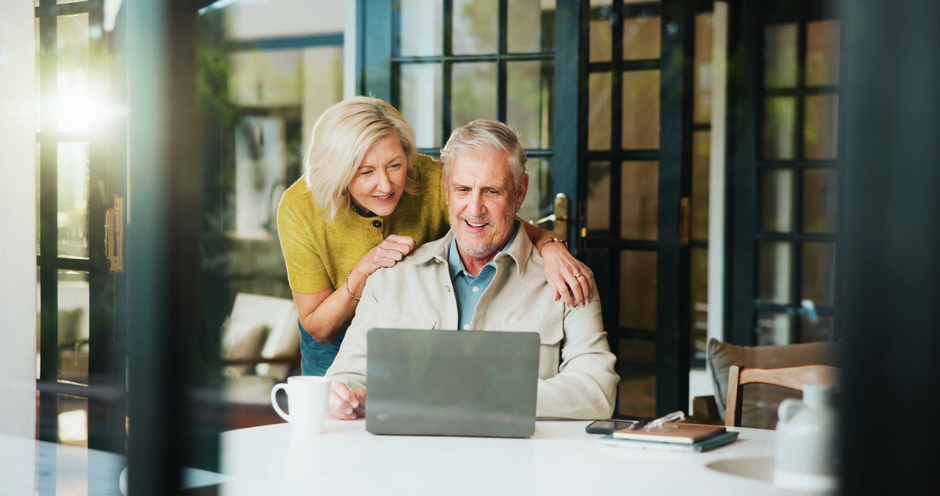 An older couple looks at a laptop together indoors. The woman has her arm around the man, smiling.