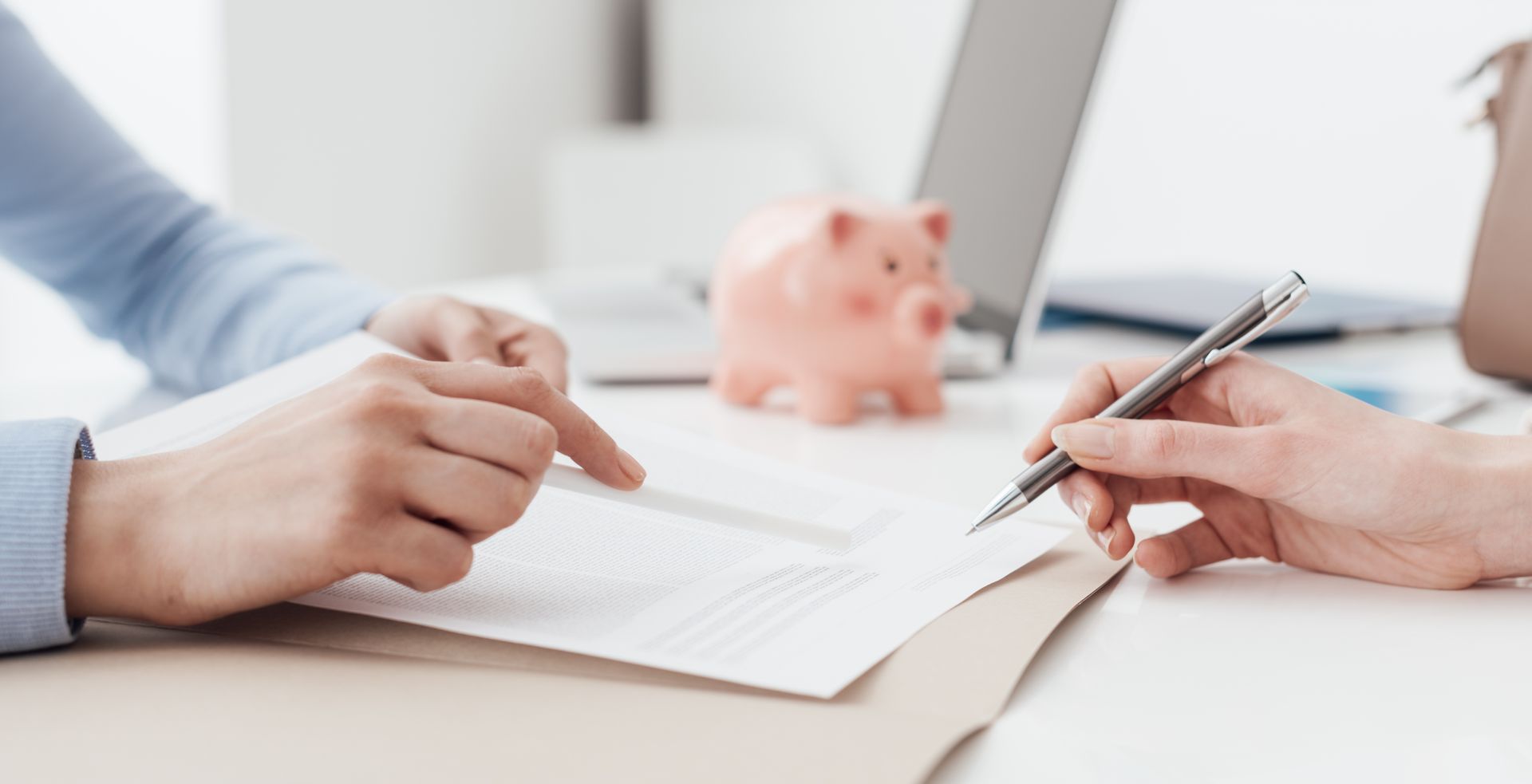 Hands pointing to a document as another hand holds a pen, near a piggy bank and laptop.