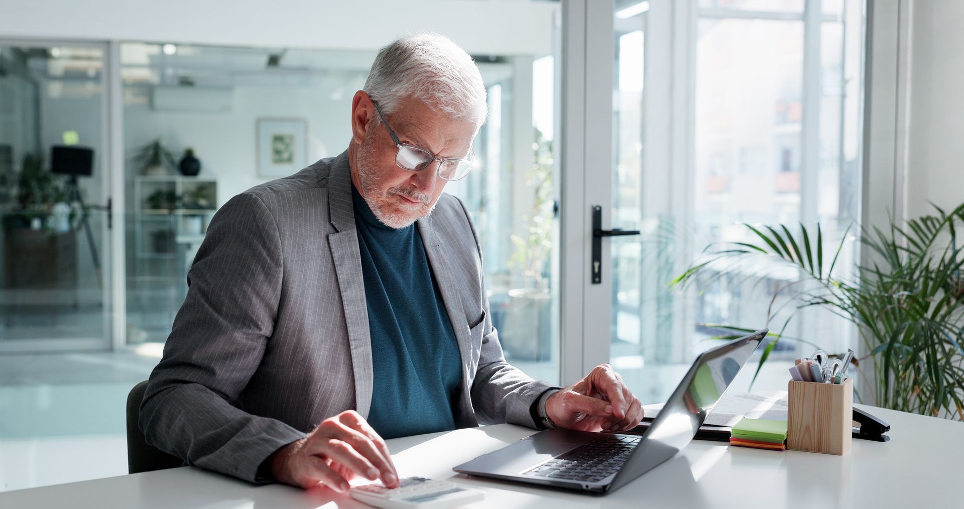 Man in a grey blazer works on a laptop at a white desk in a well-lit office.