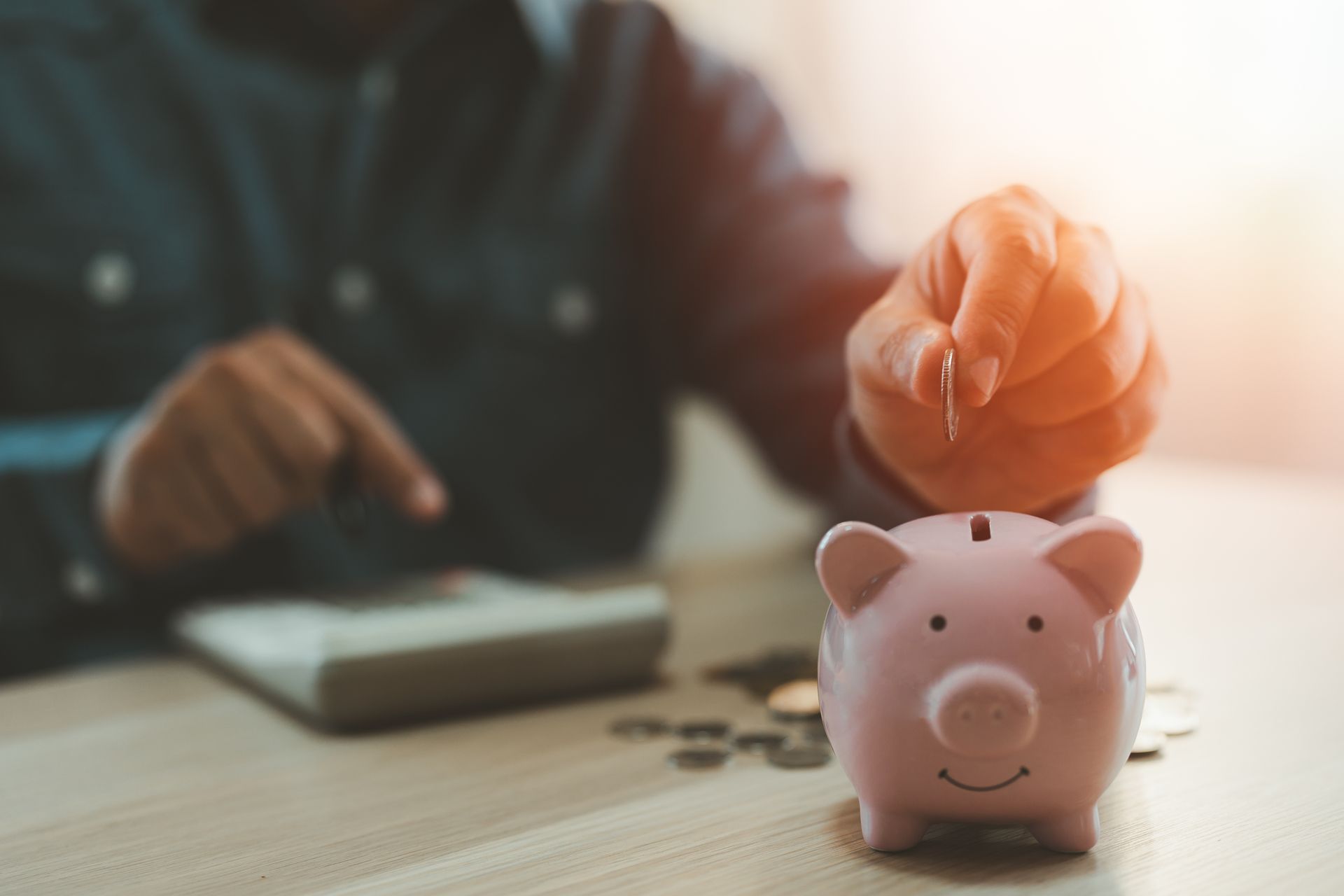 A hand dropping a coin into a pink piggy bank on a desk next to coins and a calculator.