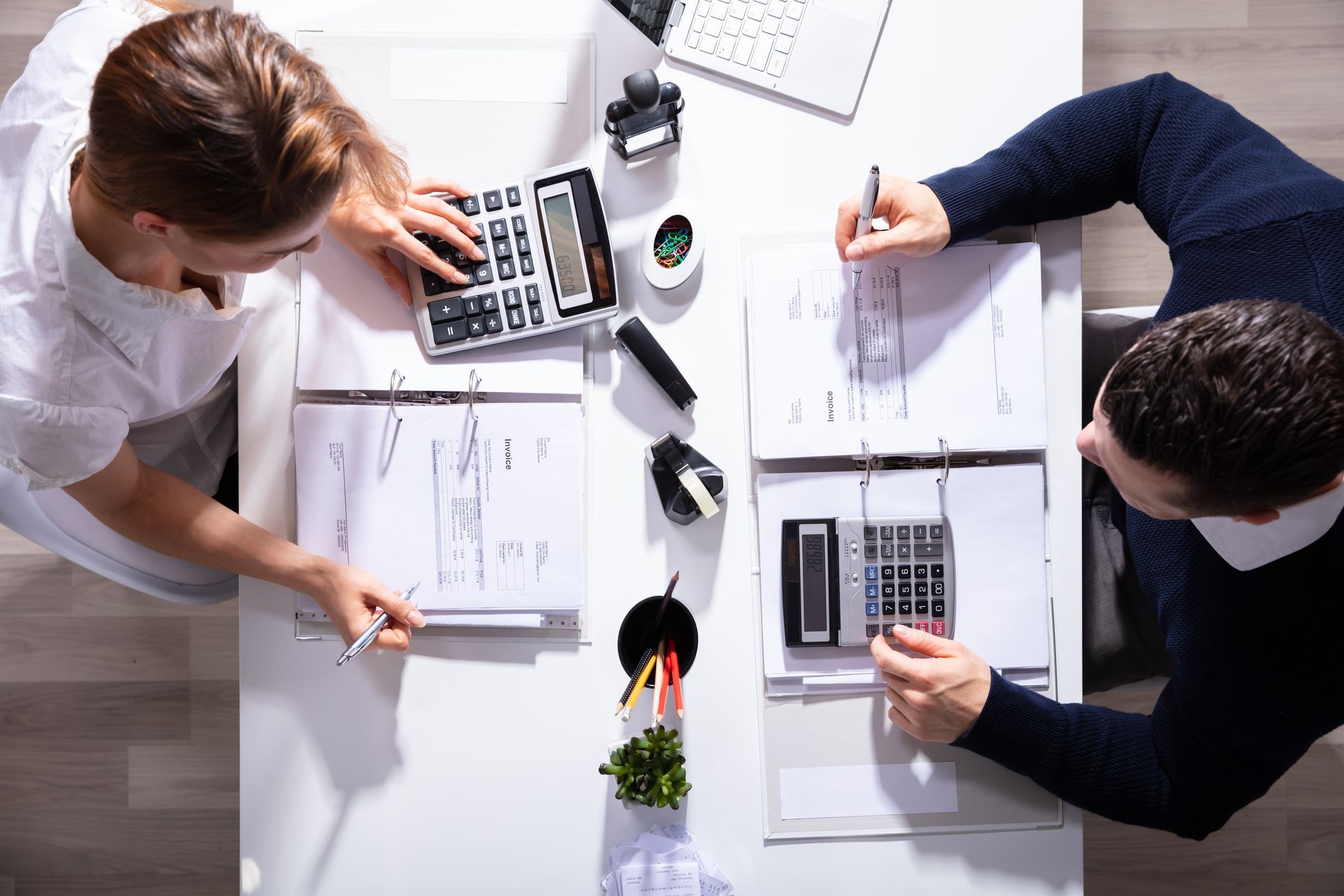 Two people at a white desk, using calculators and writing on papers. Overhead shot. Two people at a white desk, using calculators and writing on papers. Overhead shot.