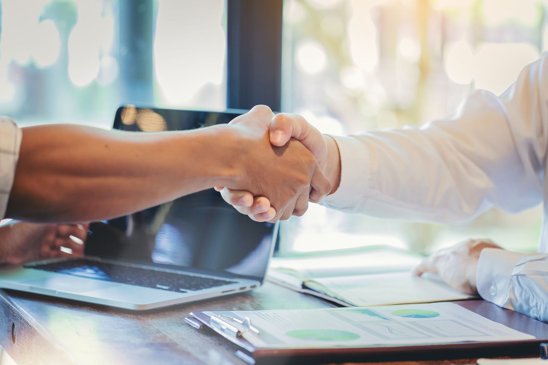 Two people shaking hands over a table with a laptop, documents, and sunlight. Two people shaking hands over a table with a laptop, documents, and sunlight.