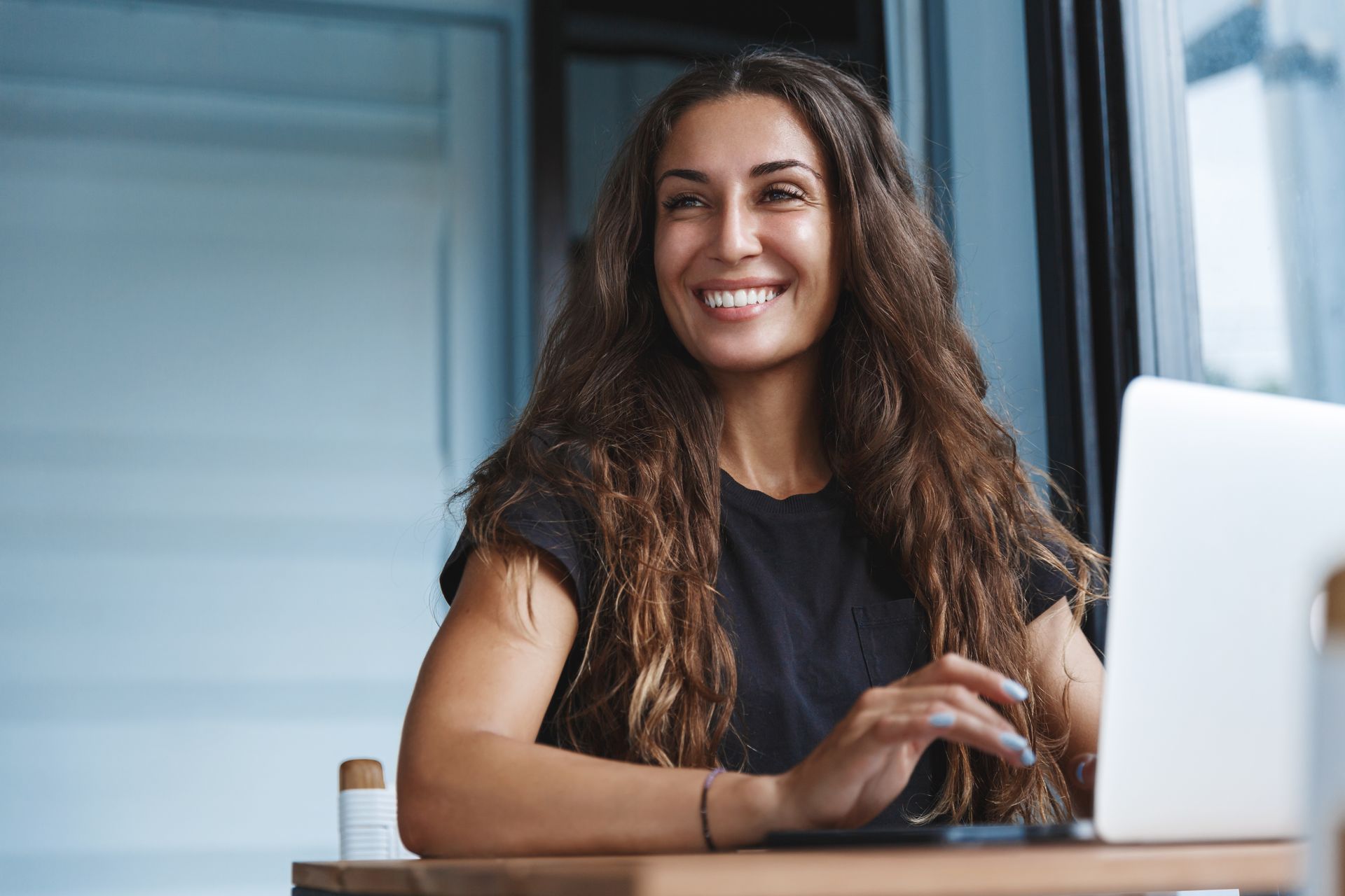 Woman with long, wavy brown hair smiles while working on a laptop at a table by a window. Woman with long, wavy brown hair smiles while working on a laptop at a table by a window.