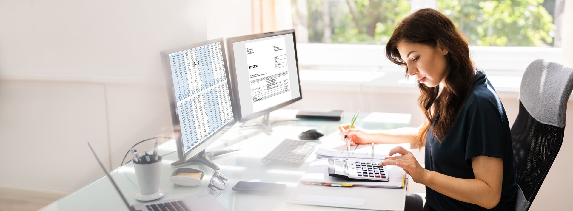 Woman at desk with dual monitors, calculating with calculator and looking at papers.