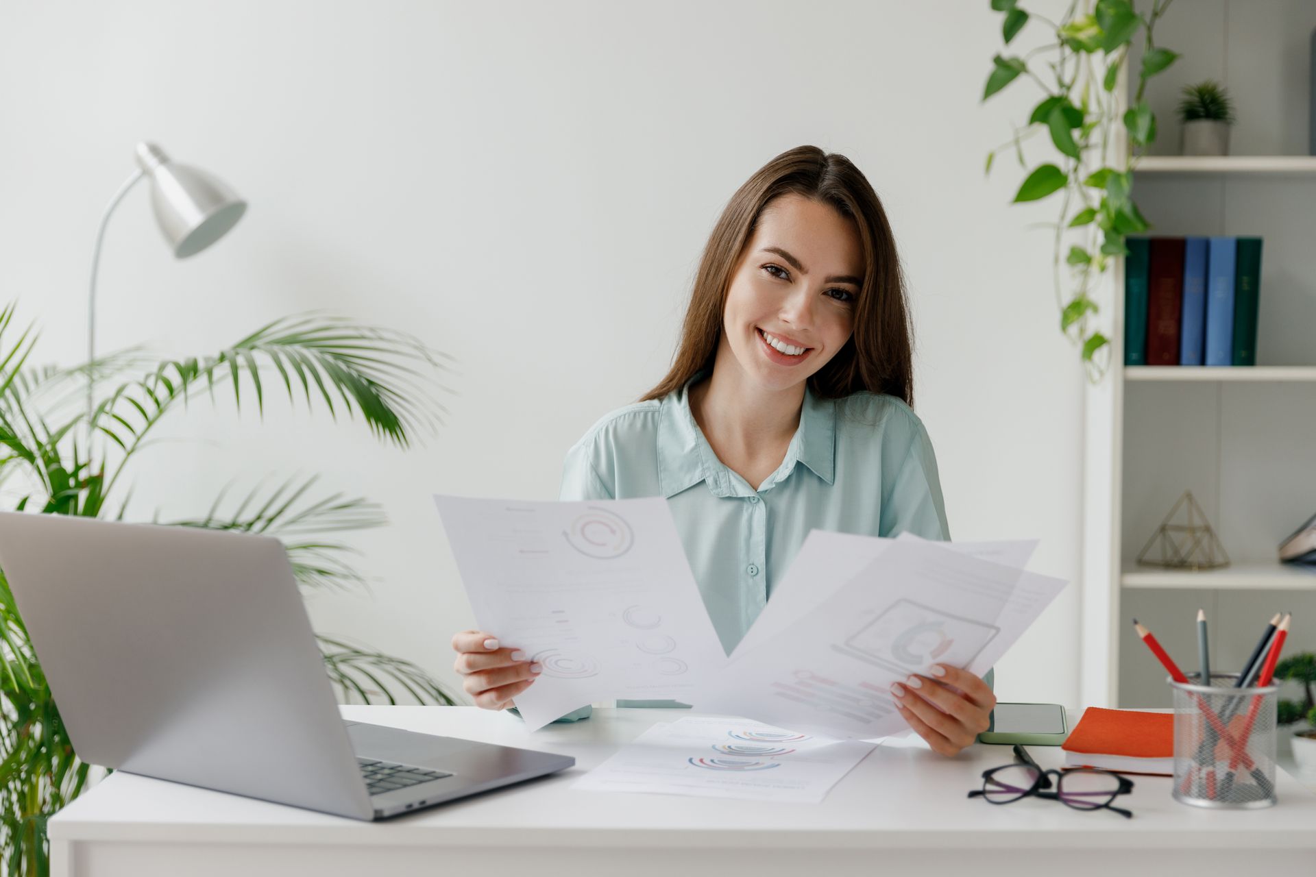 Woman smiles, holding papers, at desk with laptop, lamp, and plants.