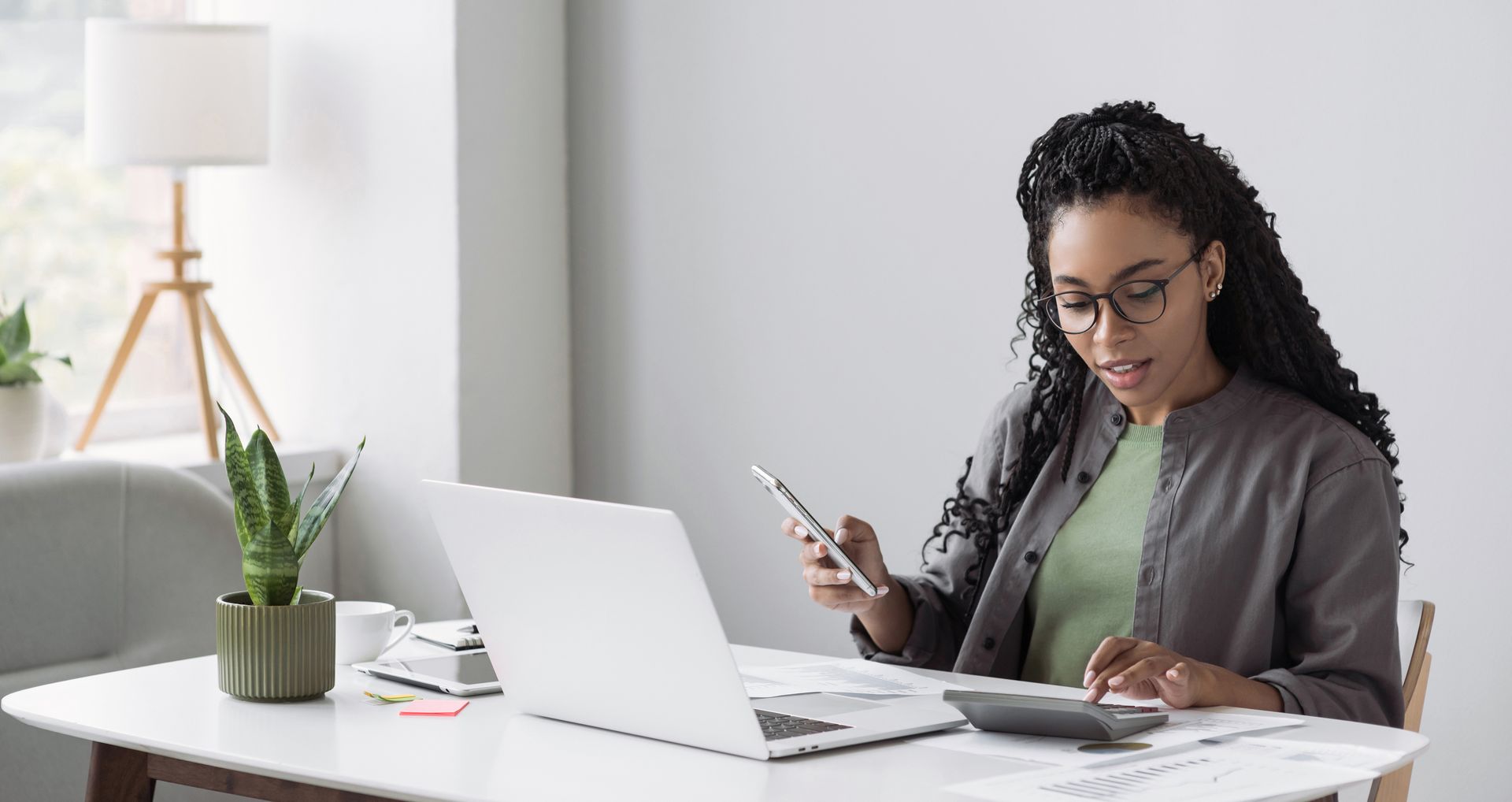 Woman wearing glasses using a calculator, laptop, and smartphone at a desk. Woman wearing glasses using a calculator, laptop, and smartphone at a desk.
