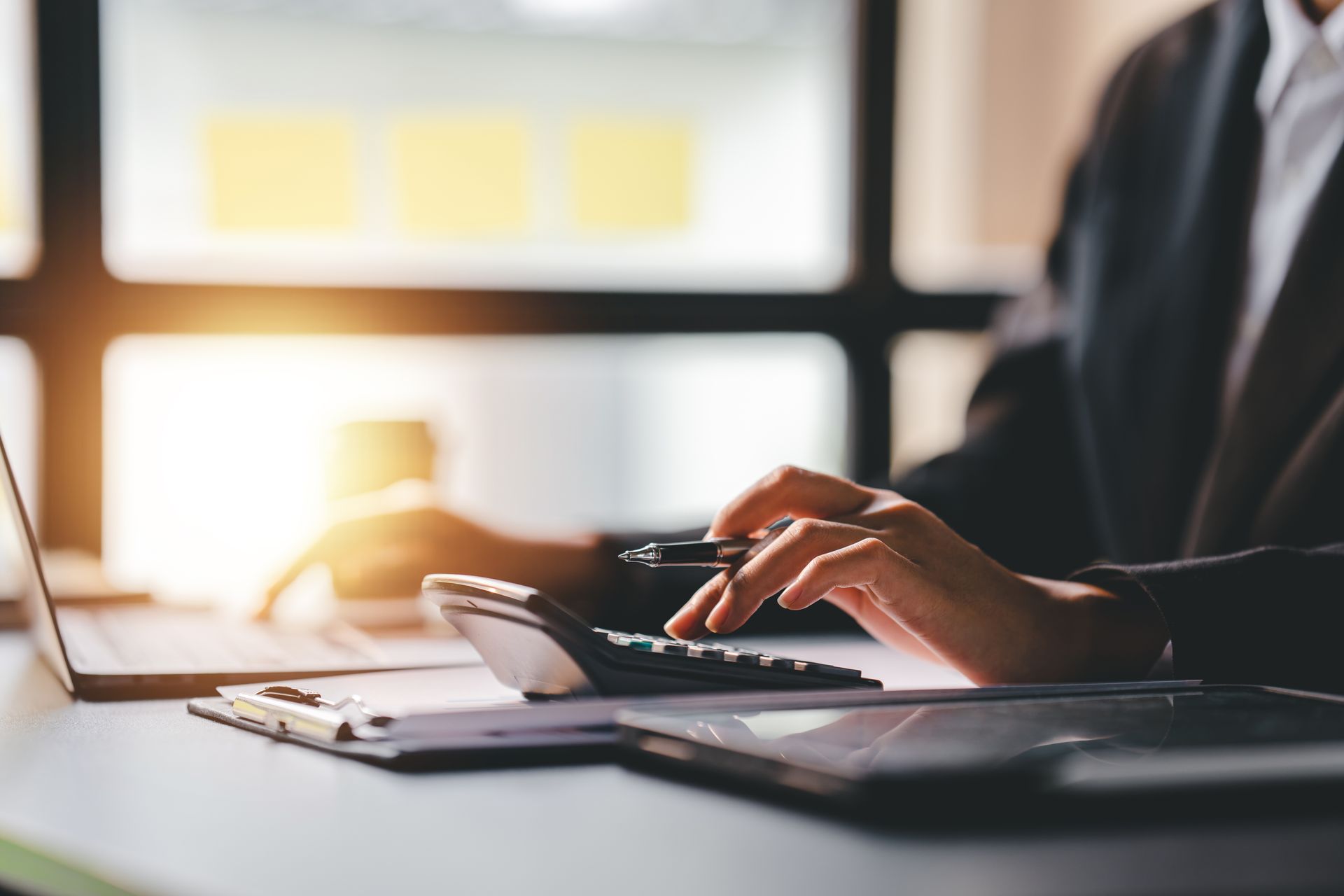 Person in a suit calculating with a calculator, using a laptop, and working on documents in an office setting.