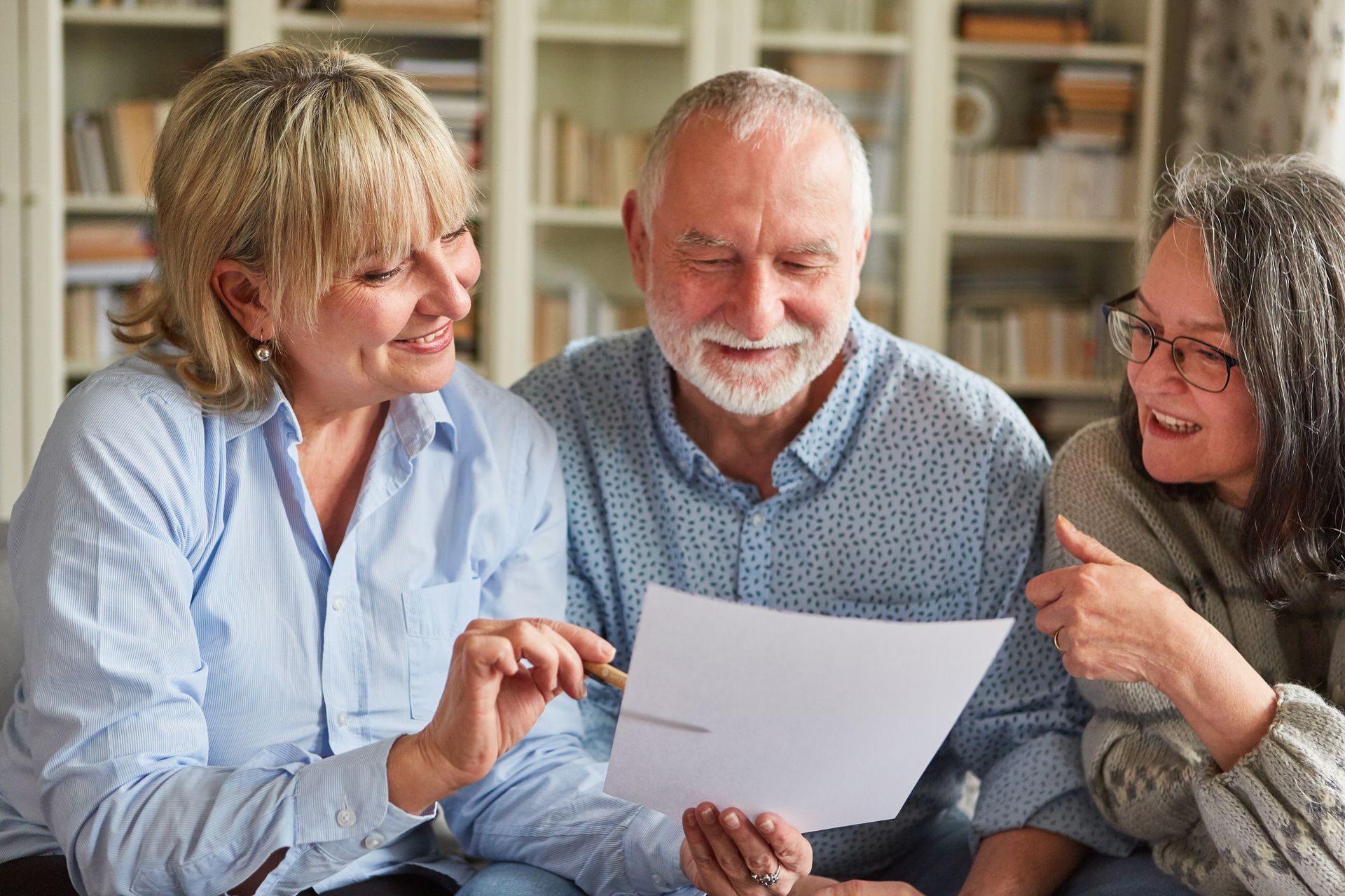 Three people looking at a document, smiling in a living room with bookshelves.