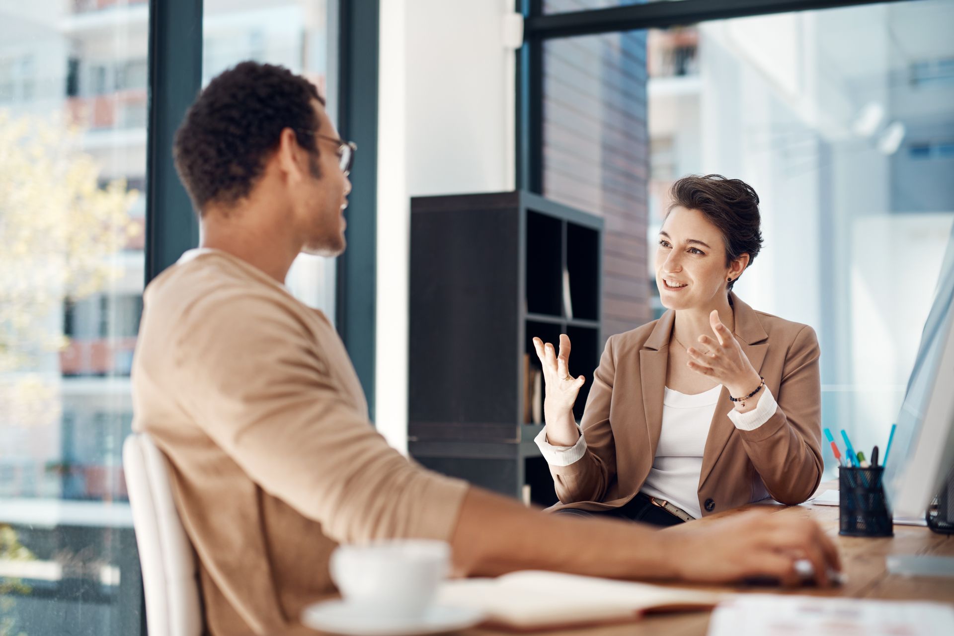 Woman gesturing, speaking with a man seated at a desk in a well-lit office. Woman gesturing, speaking with a man seated at a desk in a well-lit office.