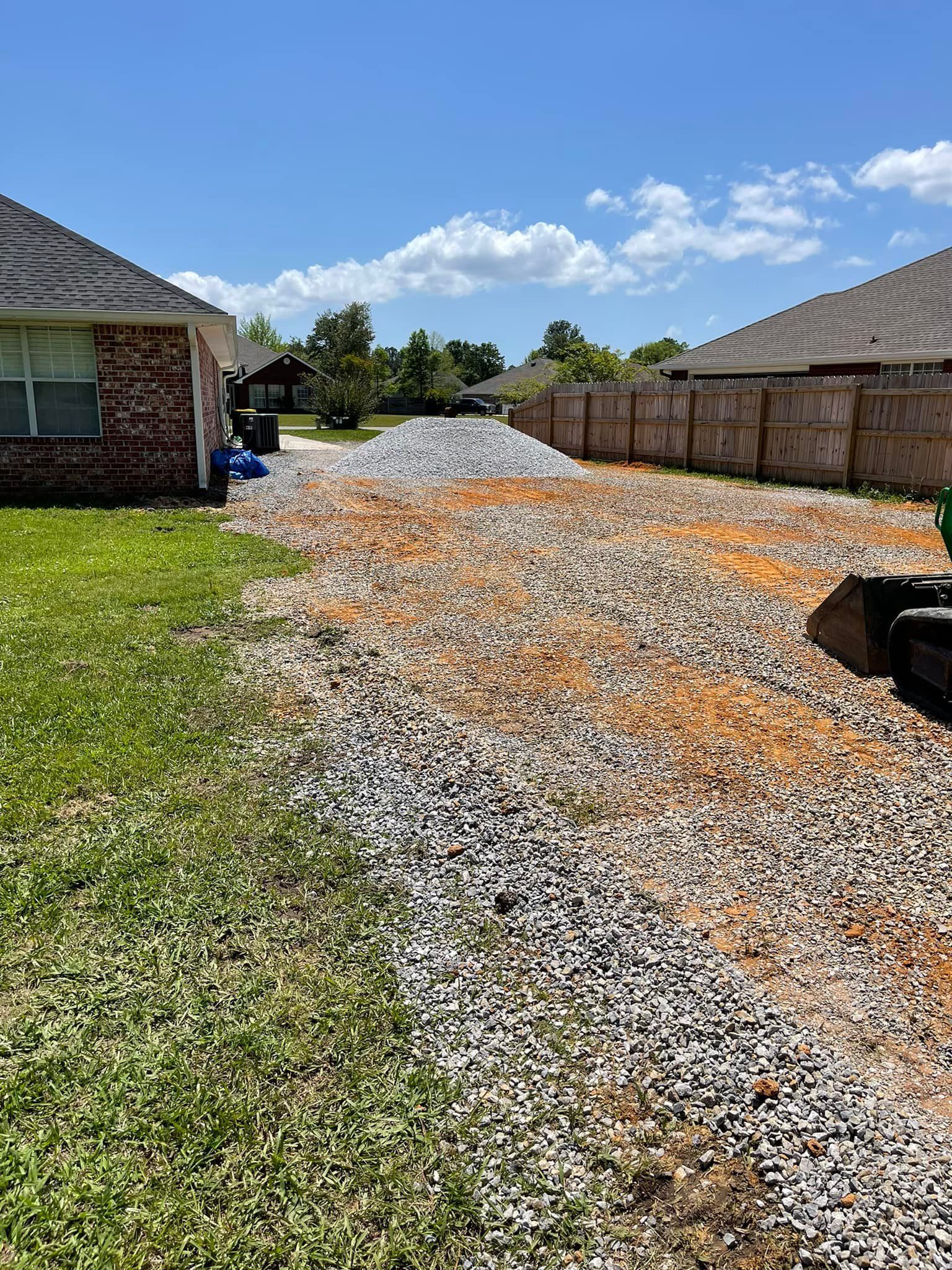 Gravel driveway under construction, with a large pile of gravel. Grass on the left, fence and houses in the background. Sunny day.