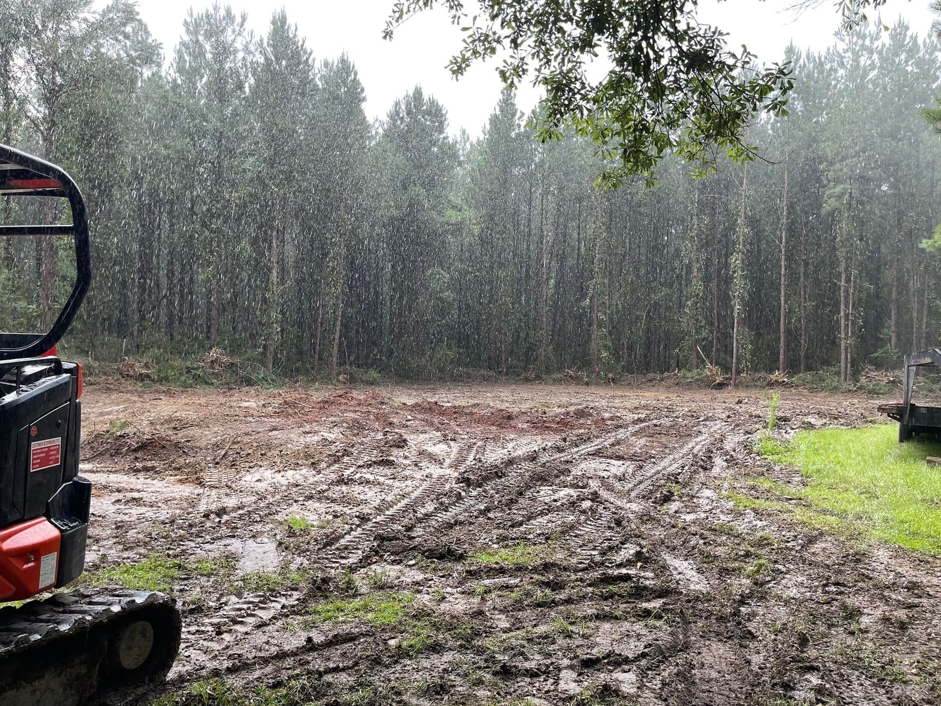 Muddy clearing in rain, with trees in background and small tractor on left.