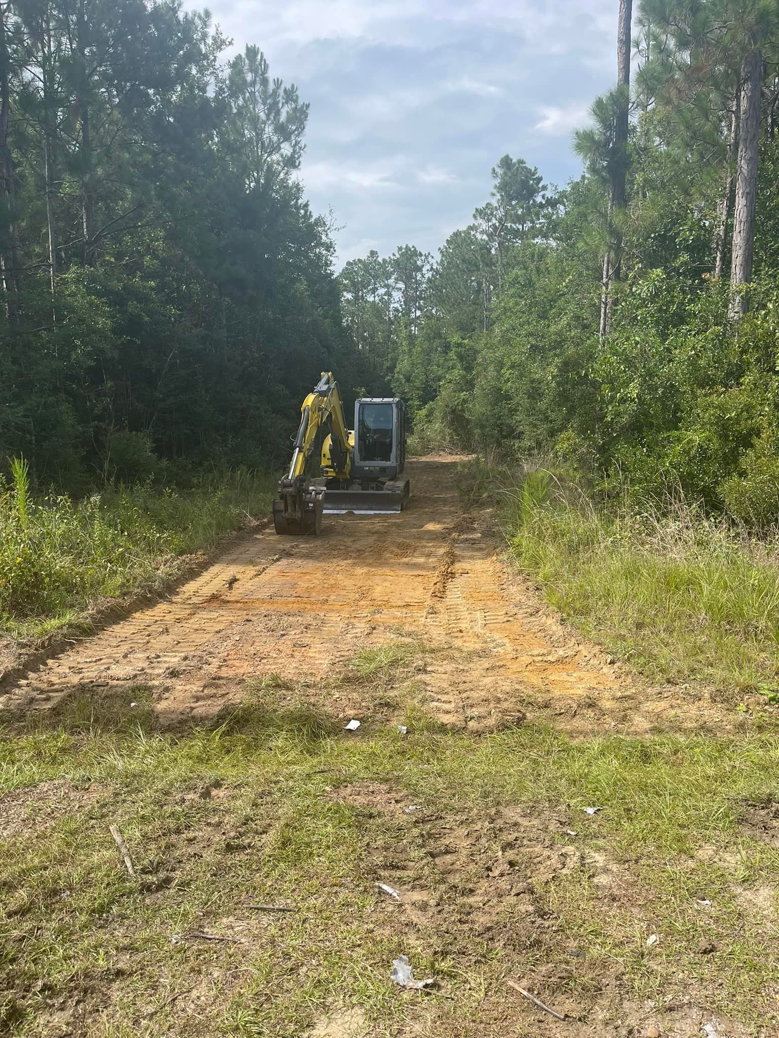 Mini excavator clearing a dirt path in a wooded area, under a cloudy sky.