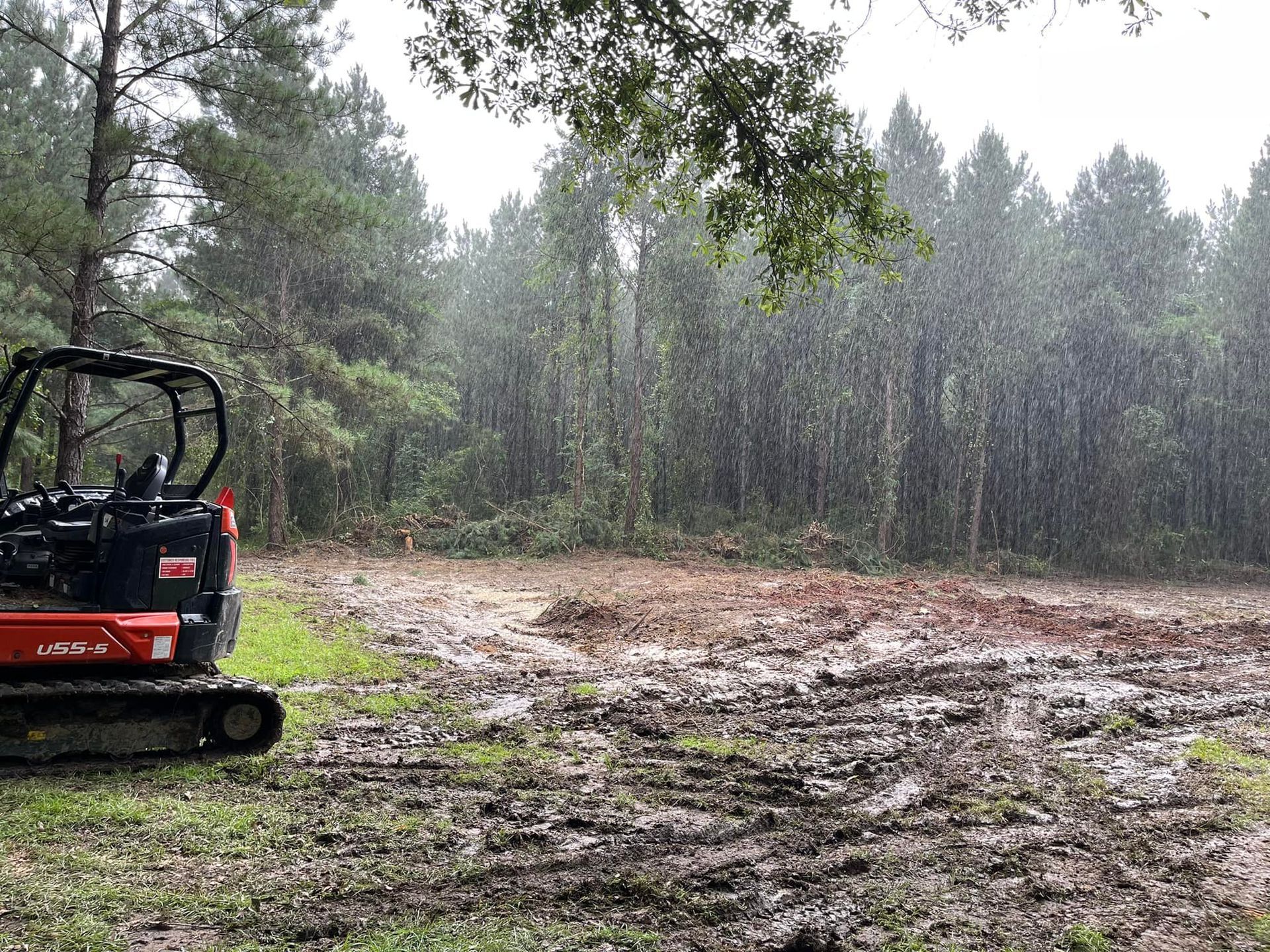 Mini-excavator sits in muddy area in the rain, clearing land. Forest in background.