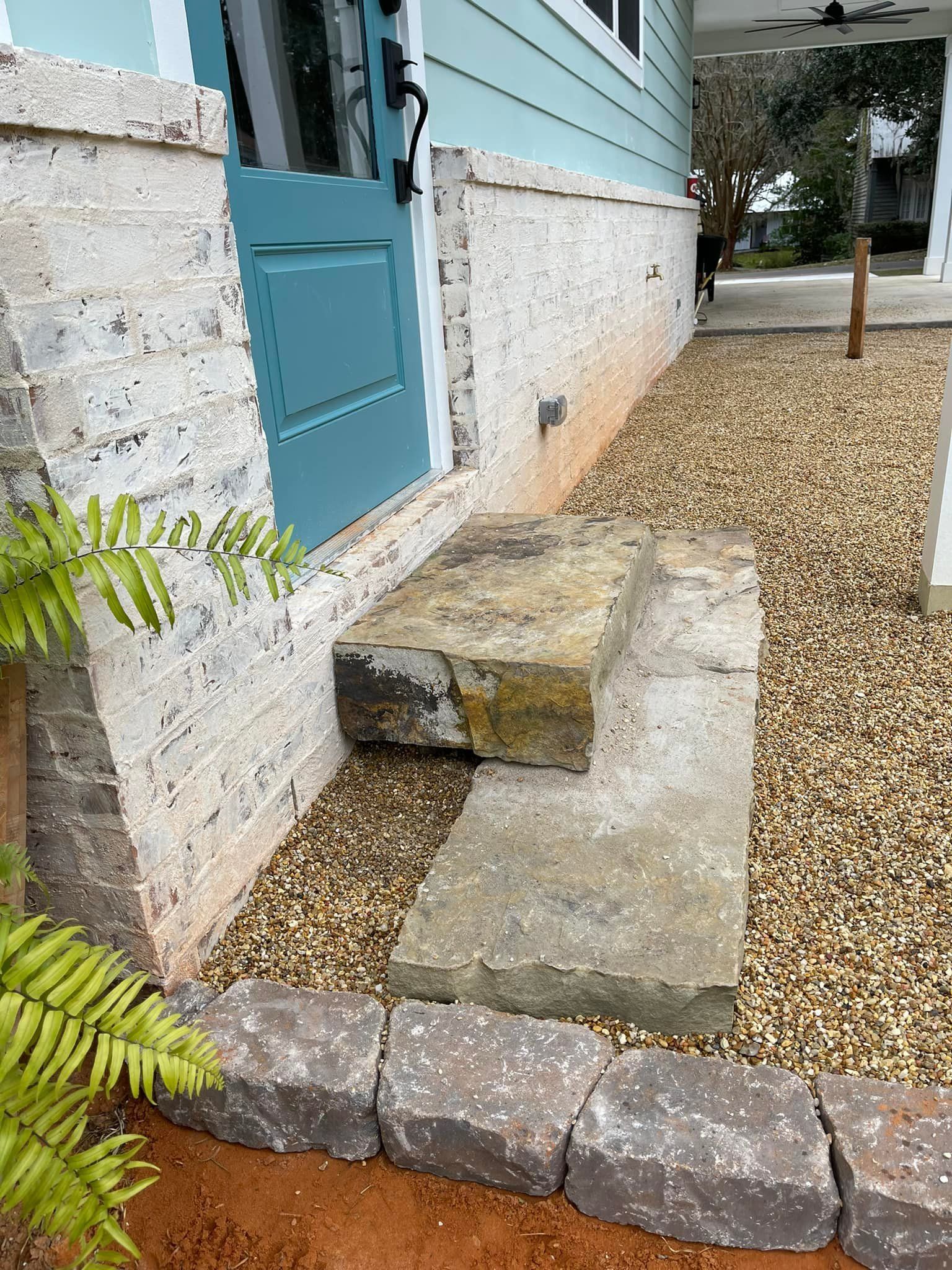 Stone steps lead to a teal door on a light blue house with a gravel pathway and brick border.