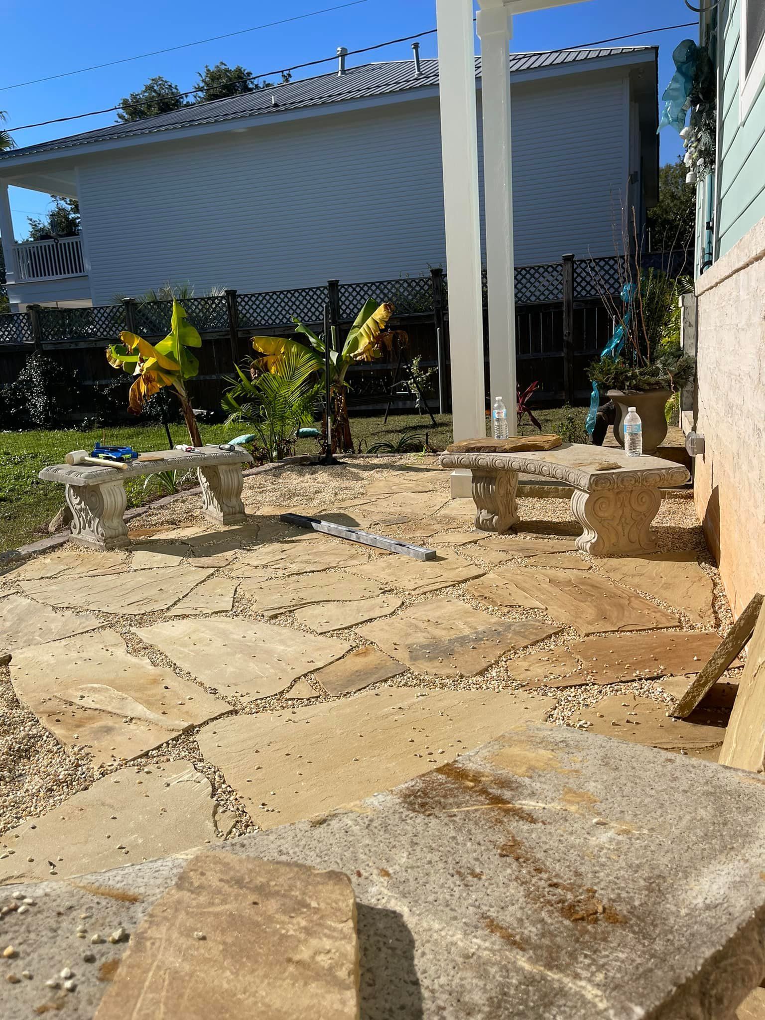 Stone patio with two benches, plants, and a white pergola, sunny day.