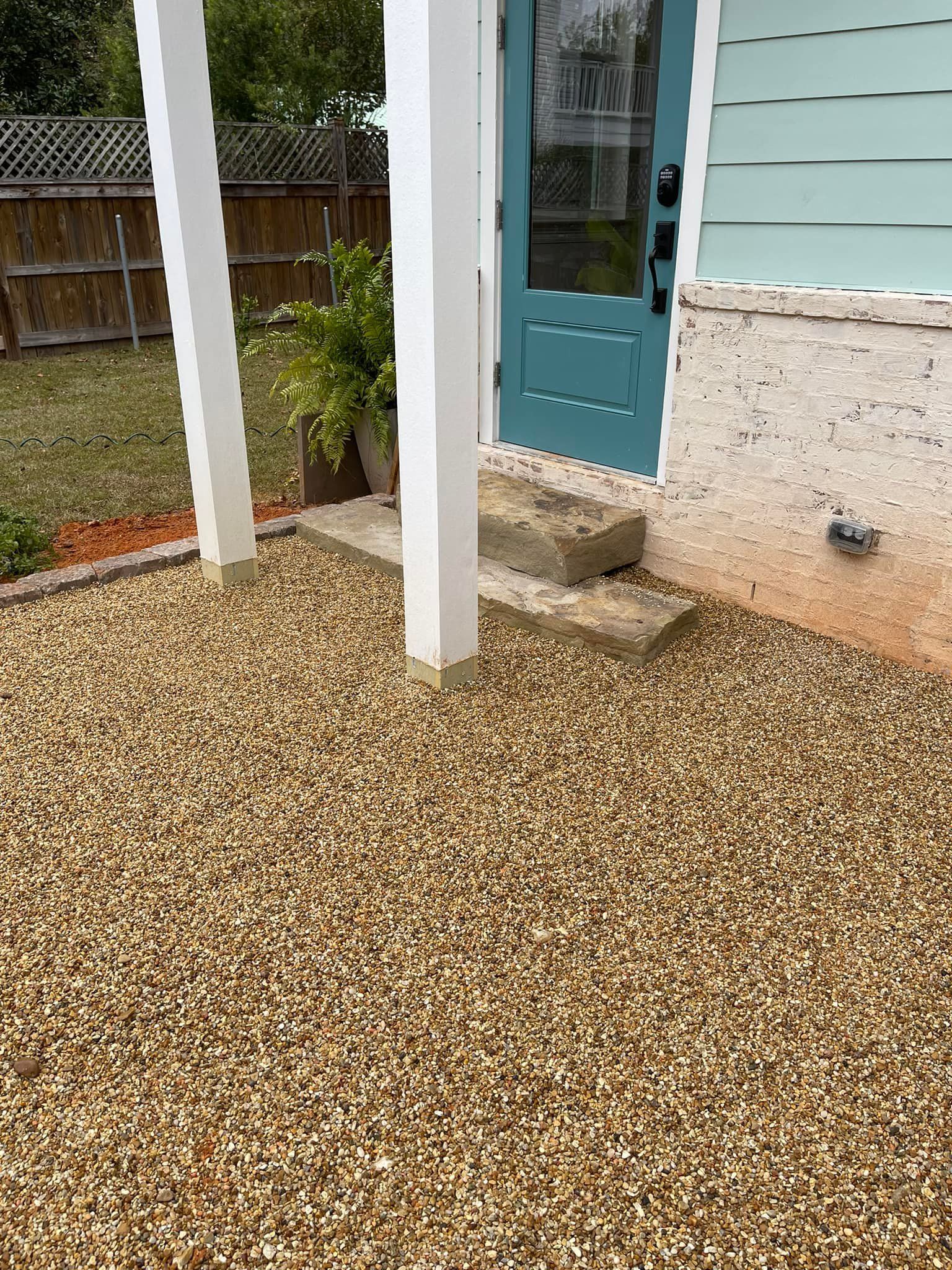 Gravel ground with a teal door, white columns, and a concrete step. A plant is in a pot beside the door.