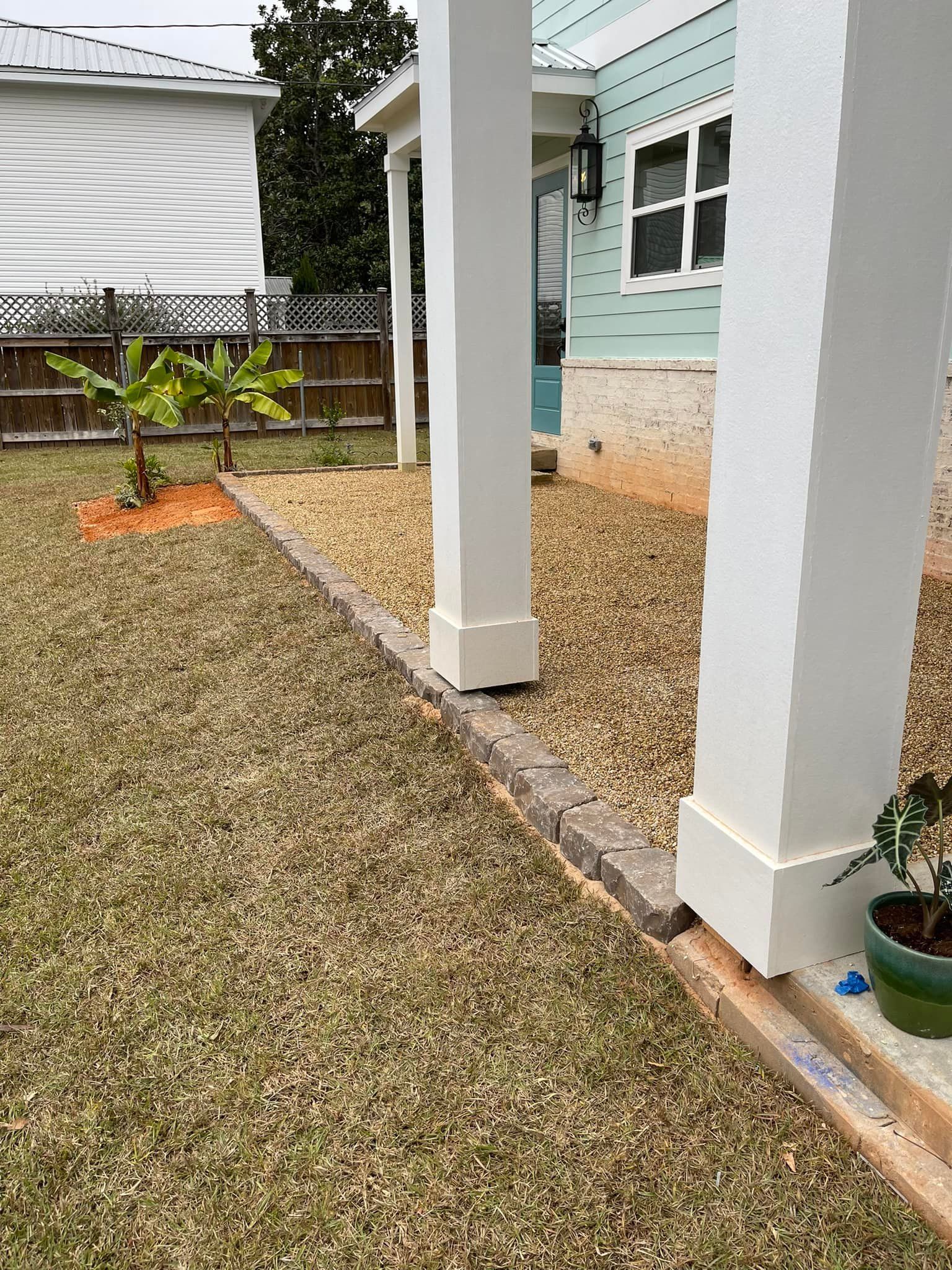 Lawn with stone border, gravel area, and porch columns. A small tree and part of a house are visible.