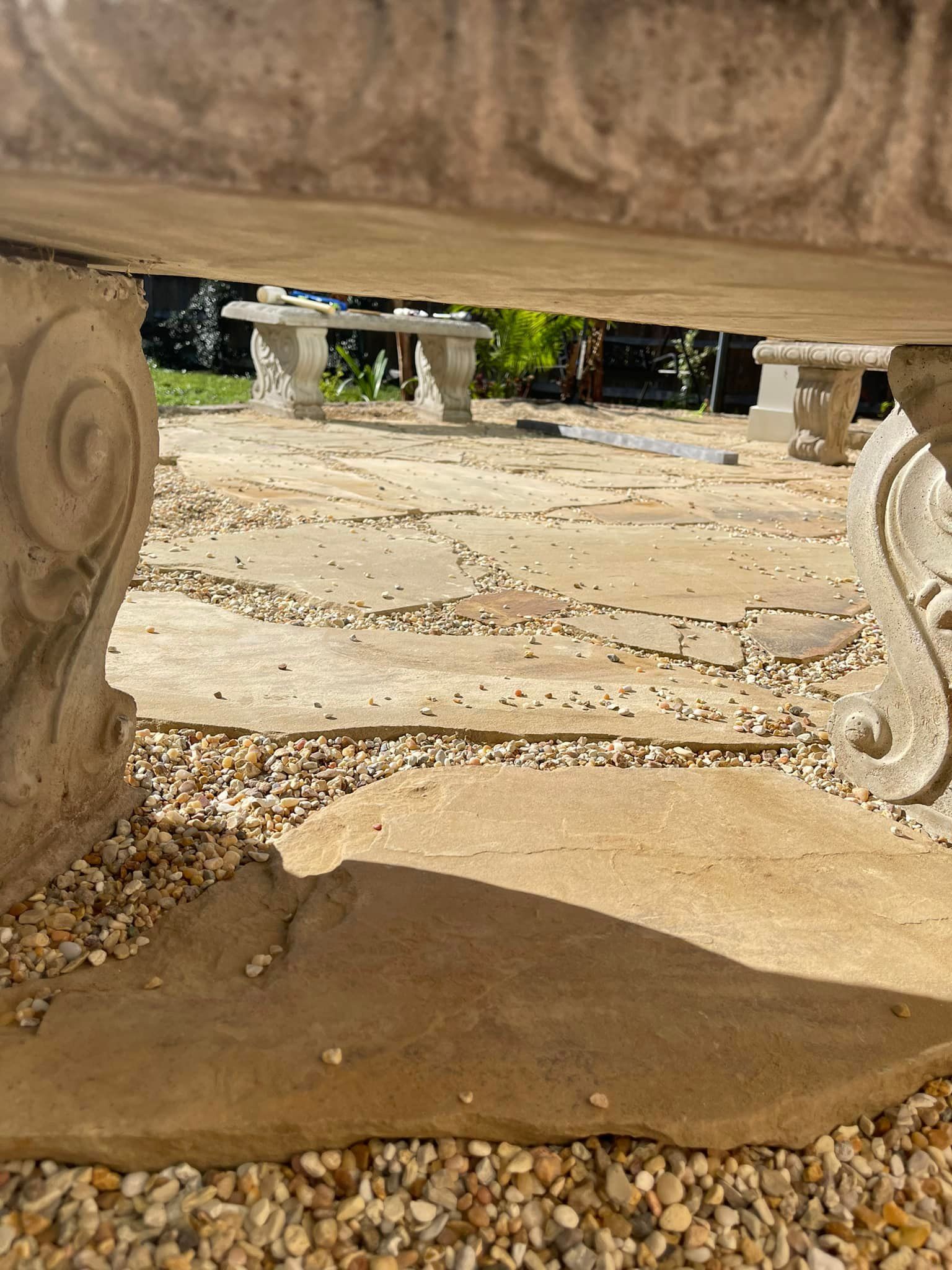 View from under a stone planter toward stone benches on a gravel patio. Sunny outdoor setting.