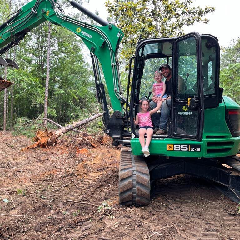 A man and two children in a green excavator outdoors, smiling. Dirt and trees in background.