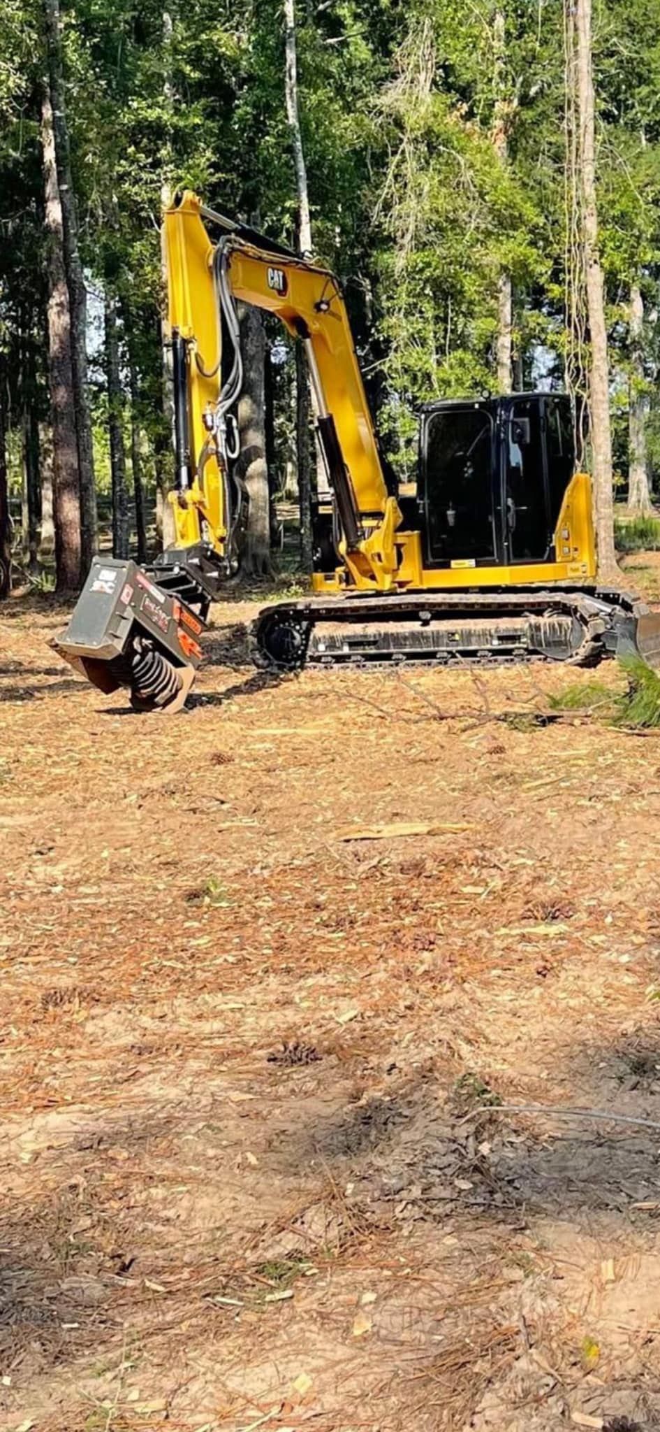 Yellow excavator with rotary cutter in a wooded area.