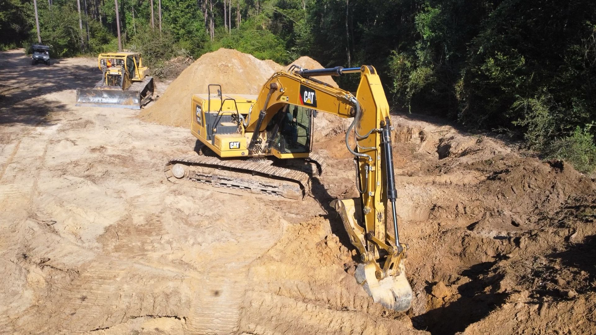 Yellow excavator digs in dirt, bulldozer in background, surrounded by trees.