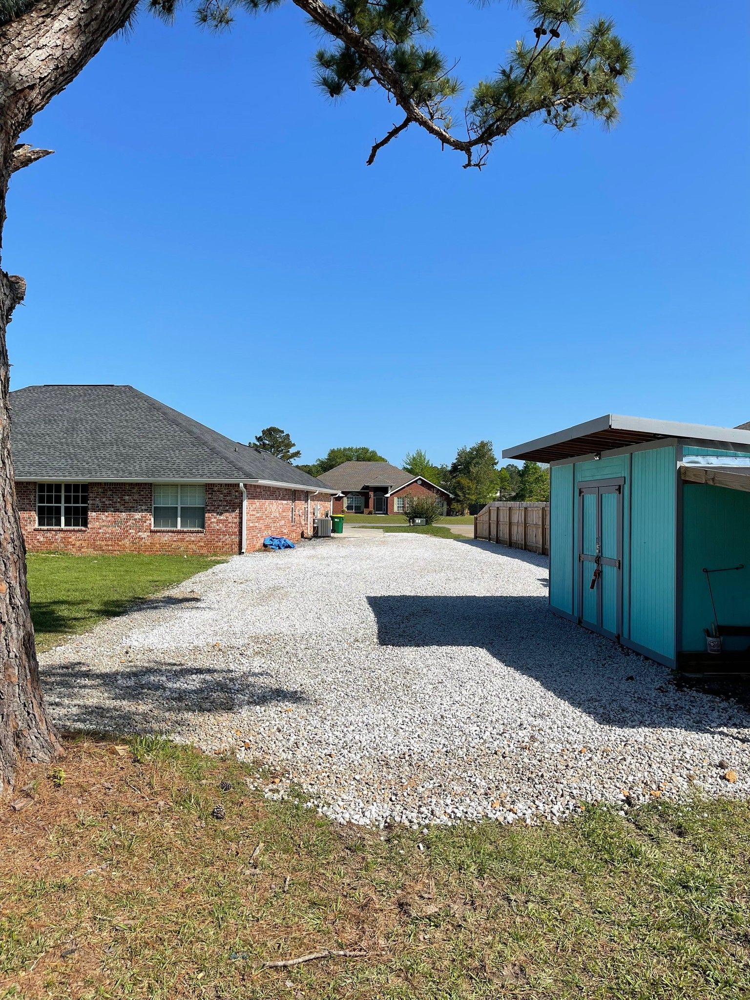 Gravel driveway between houses under a bright blue sky. A teal shed sits on the right.