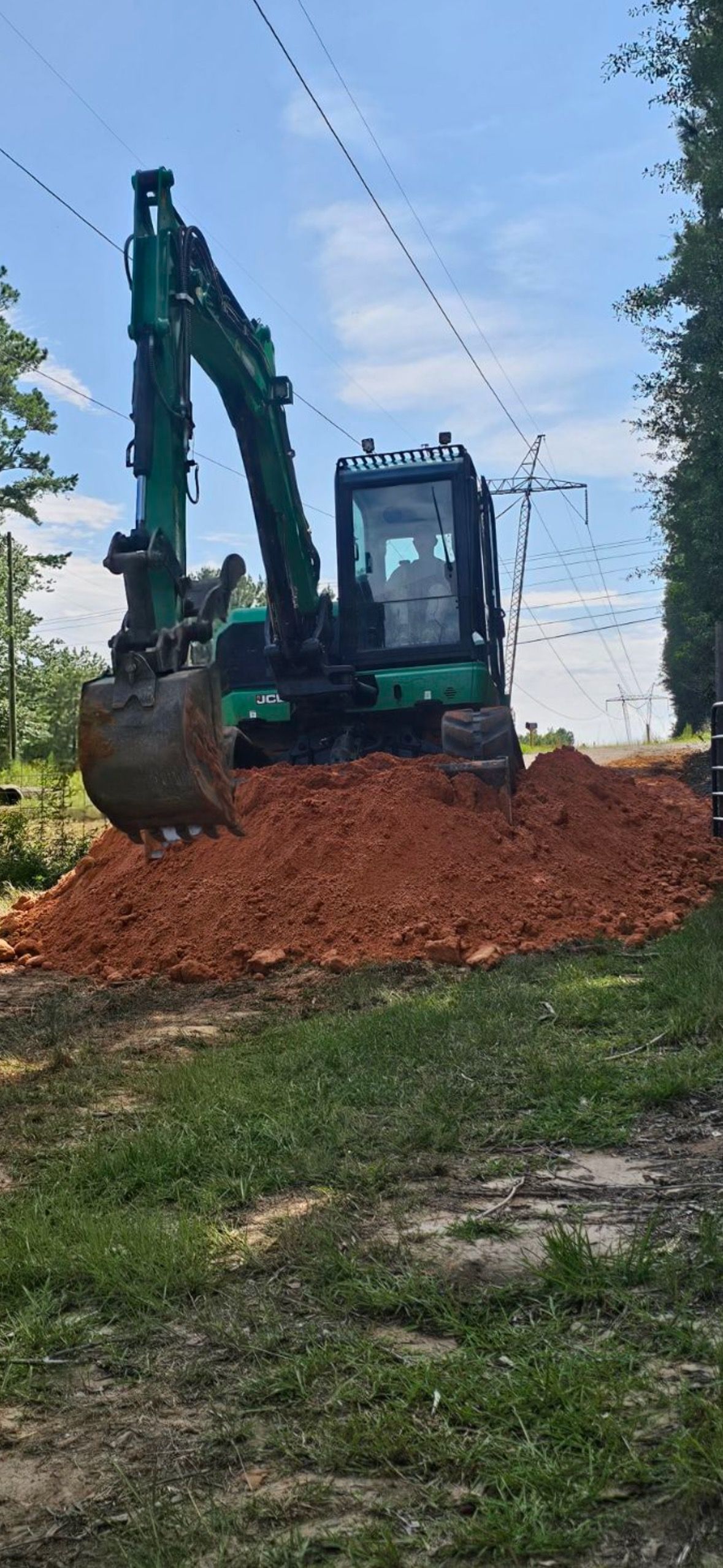 Green excavator on a pile of red dirt, under power lines.