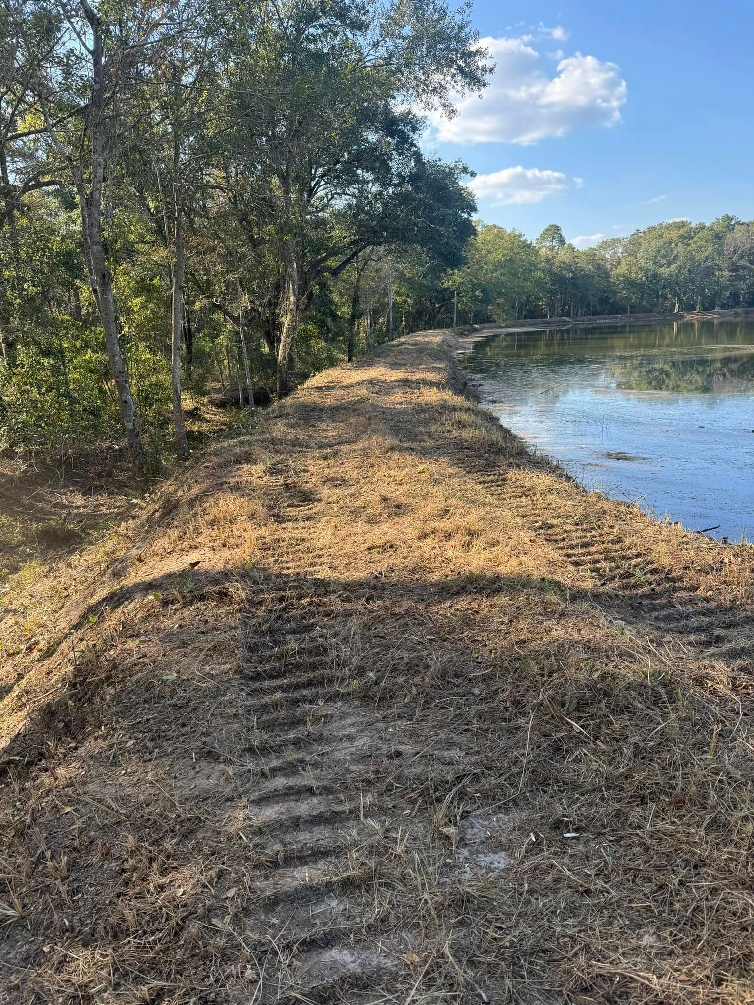 Earthen dam covered in straw, next to a lake, with trees in the background under a blue sky.