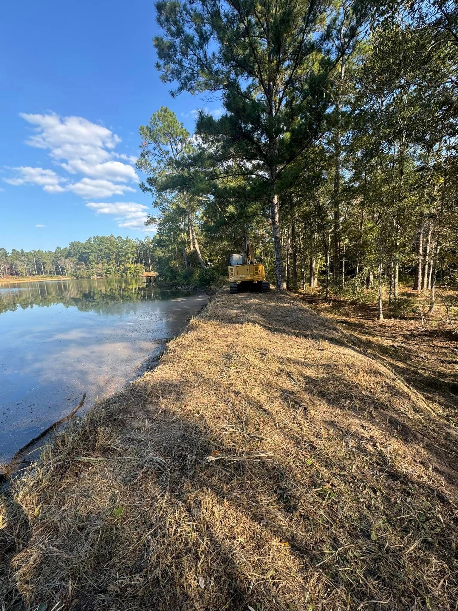 A yellow bulldozer works along the edge of a lake, clearing land near tall trees under a blue sky.