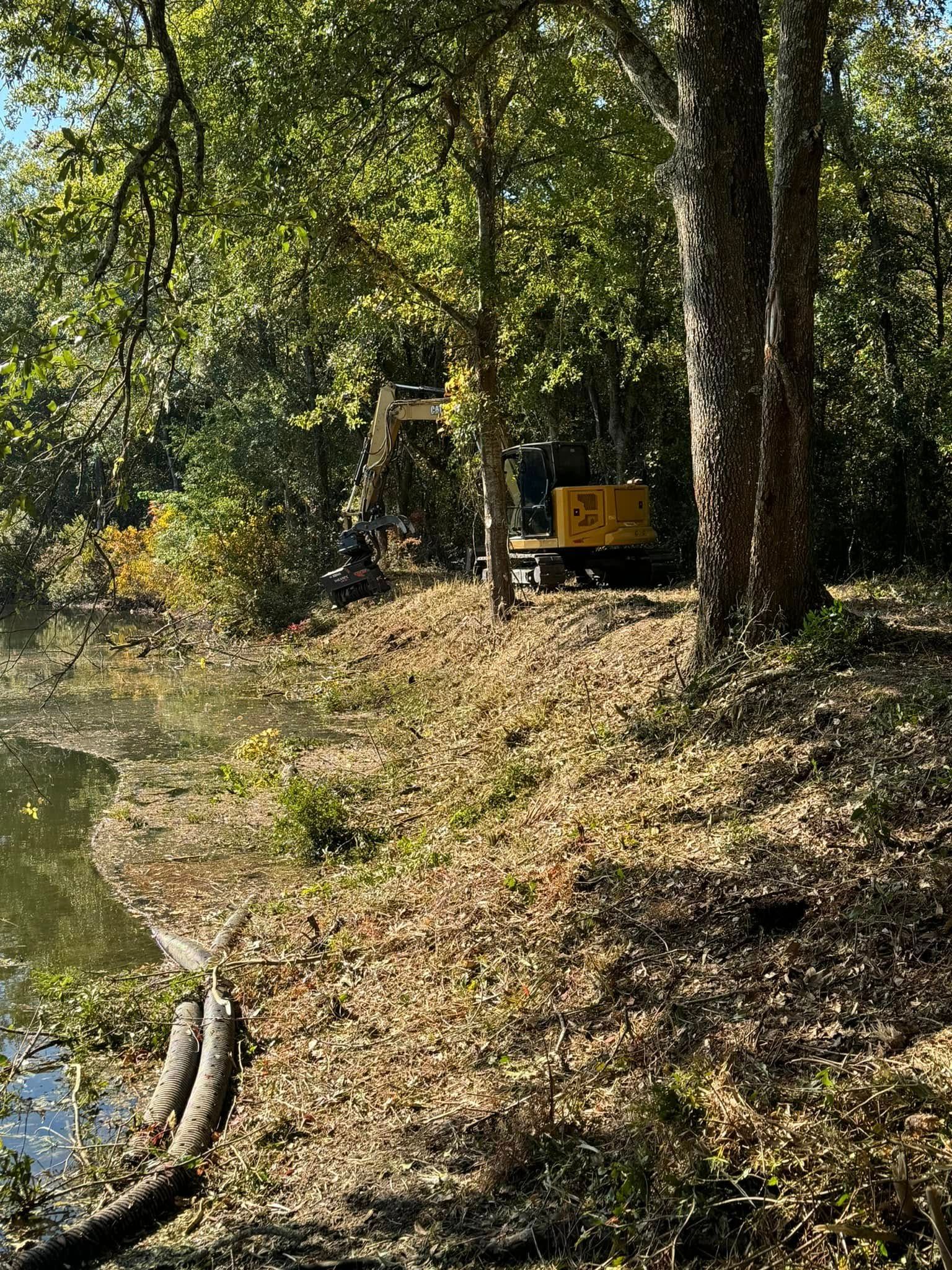 Yellow excavator on the edge of a pond, working near trees and brush.