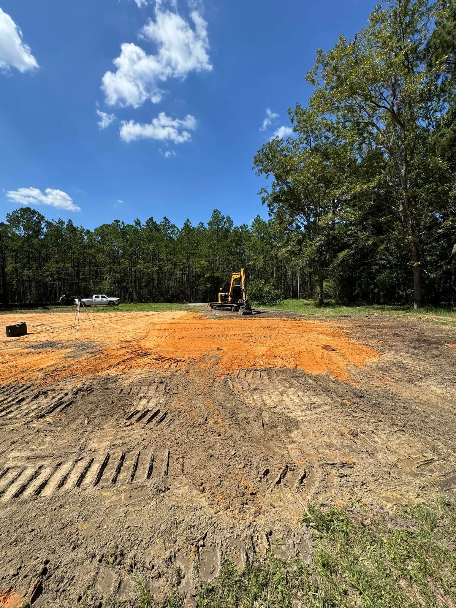 Construction site with a small excavator on cleared land, surrounded by trees and a blue sky.