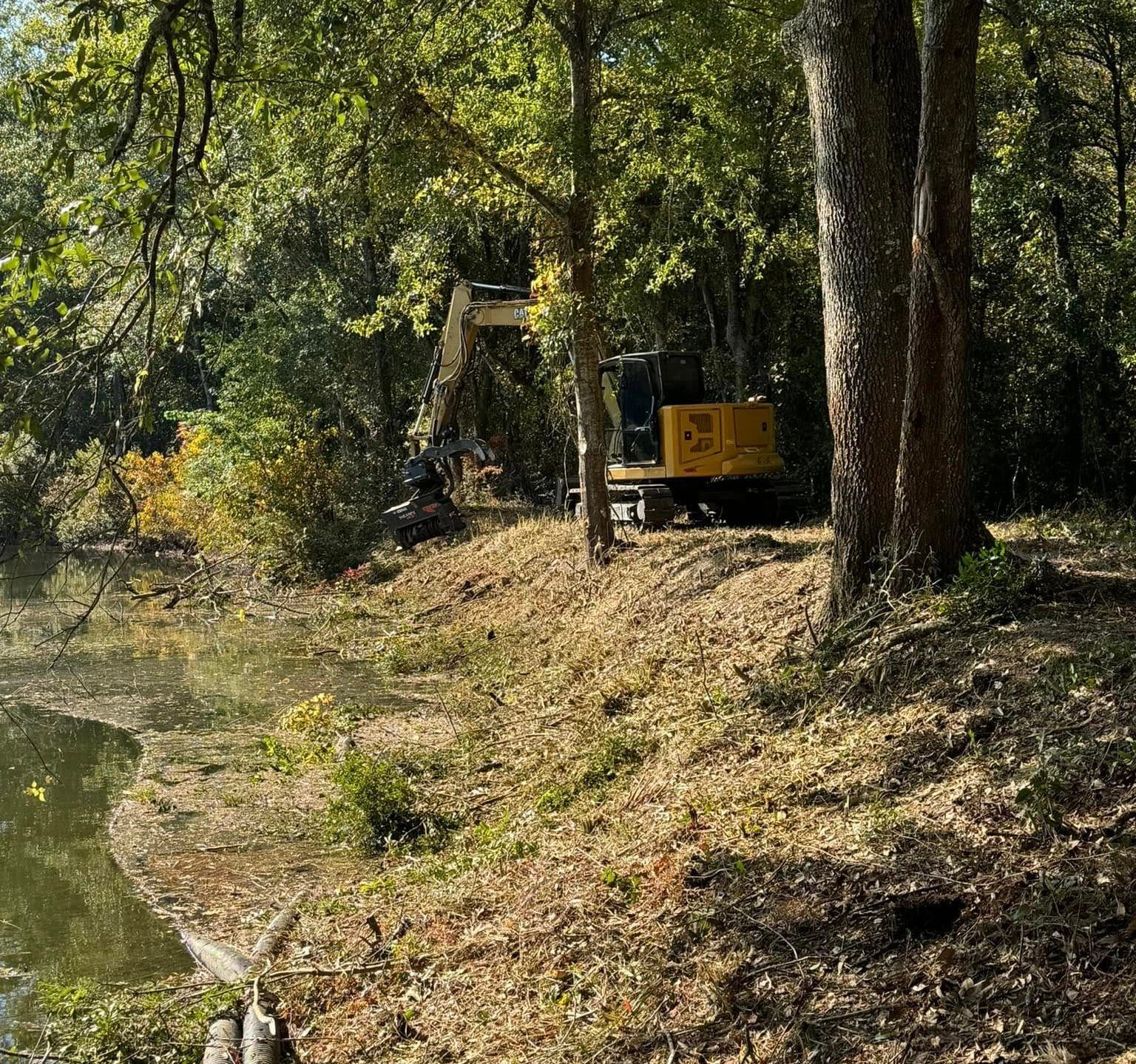 Excavator on a hill near a pond, trees surround the construction.
