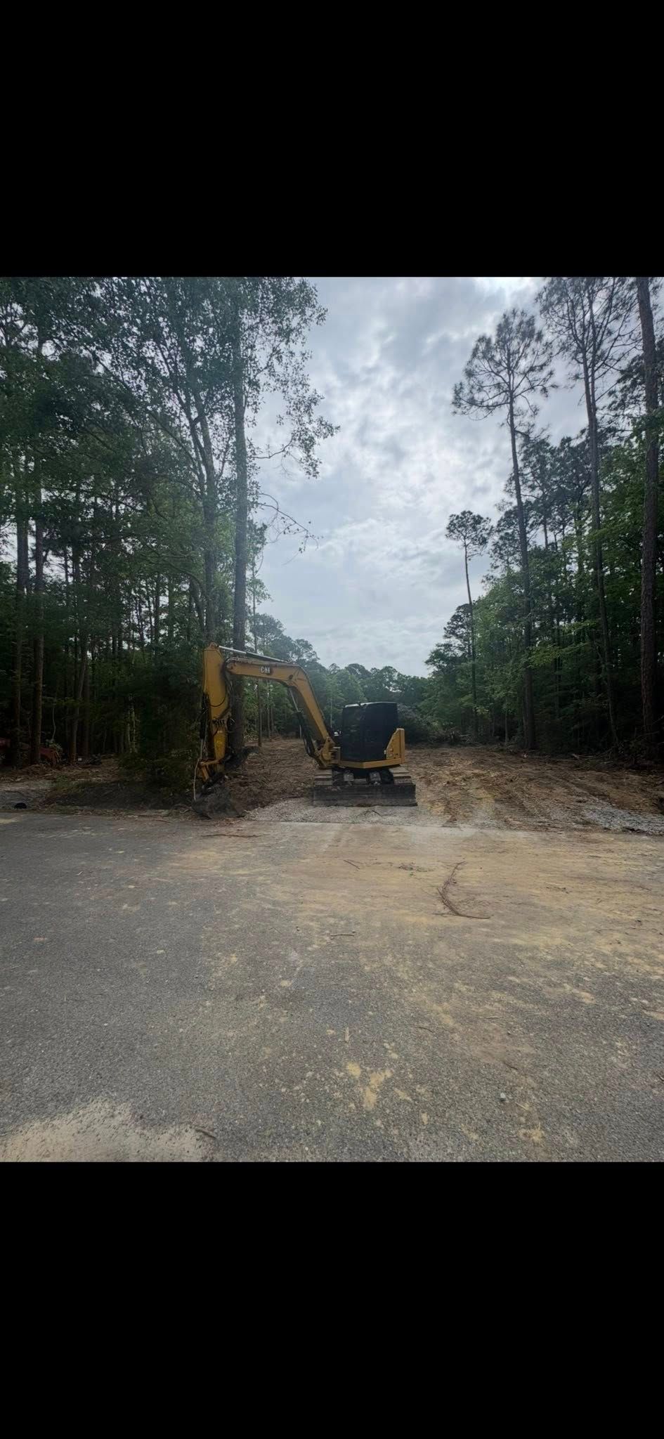 An excavator clearing a gravel road through a wooded area. Cloudy sky overhead.