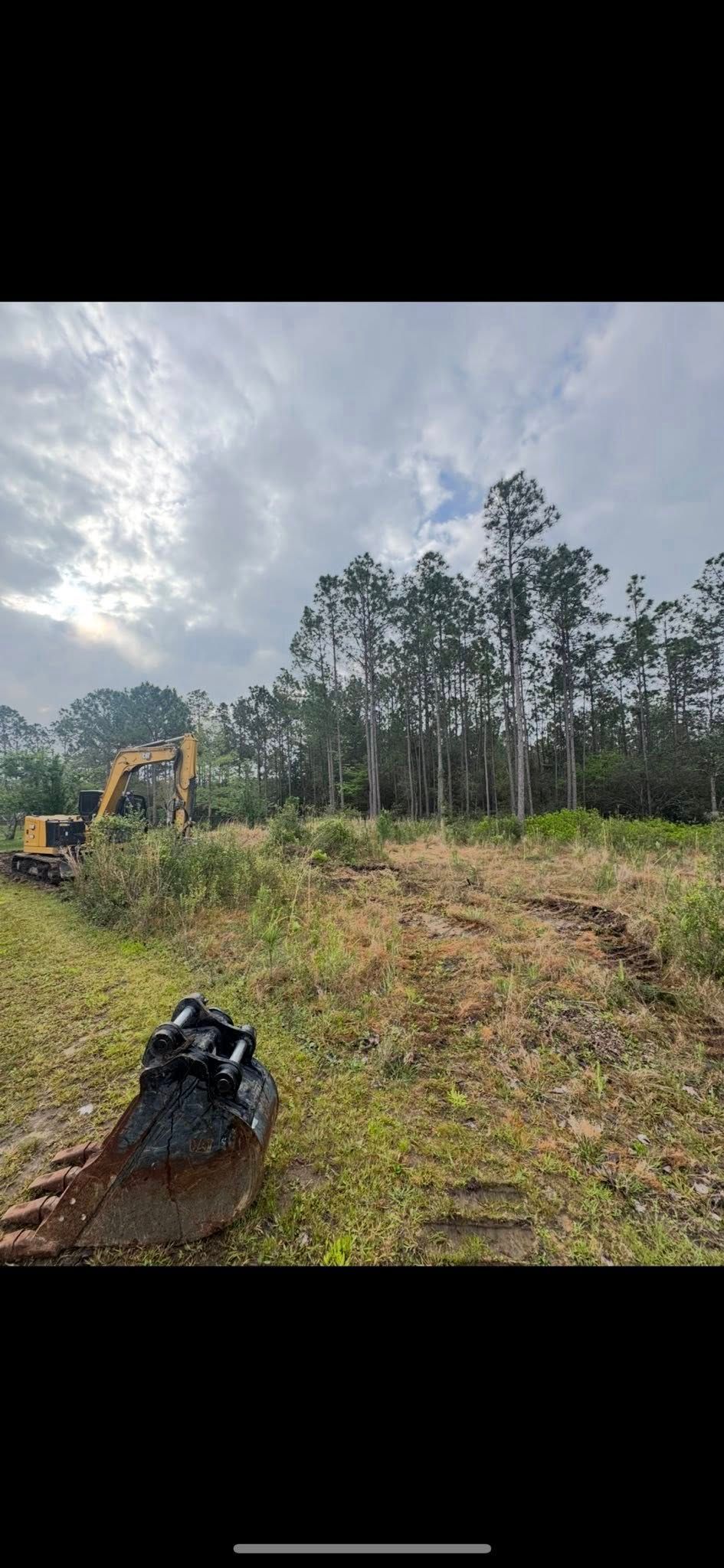 A construction site with a yellow excavator and trees, under a cloudy sky.