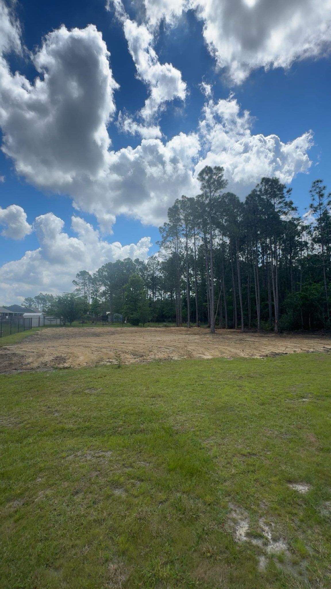 Grassy field with cleared dirt patch, trees in the background, blue sky with clouds.