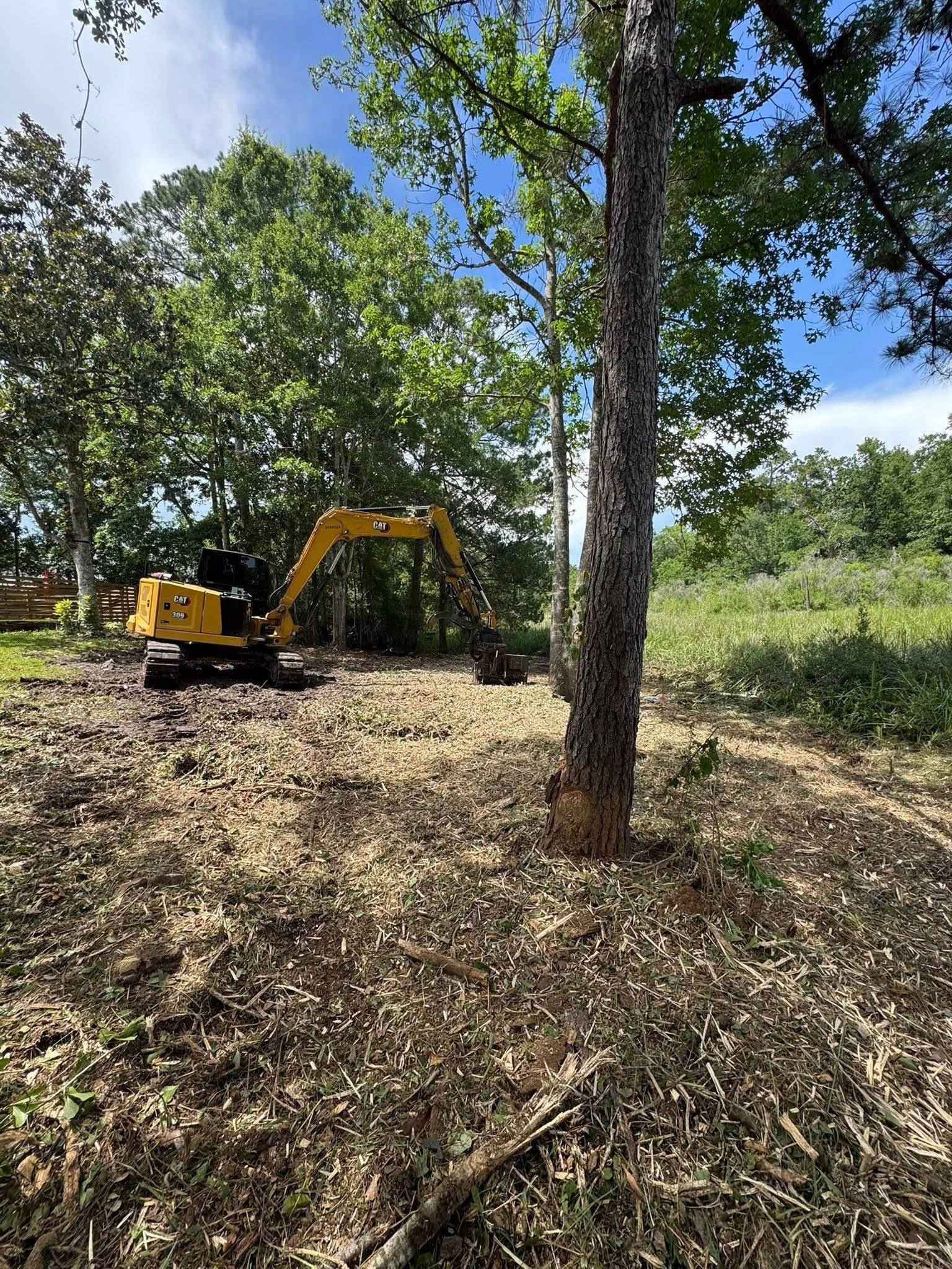 Yellow excavator clearing brush from a wooded area under a blue sky.