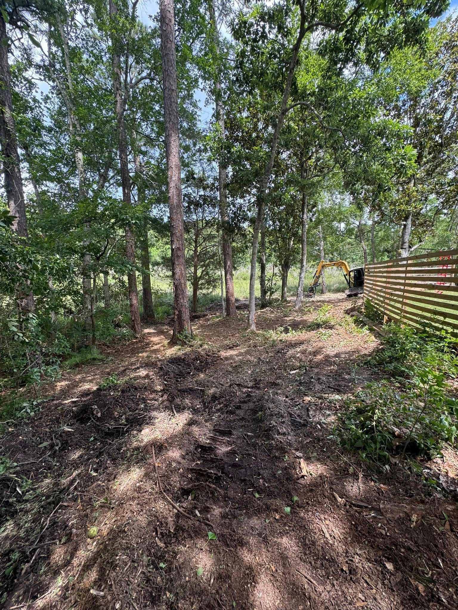 Dirt patch in wooded area with trees, a fence, and an excavator in the background.