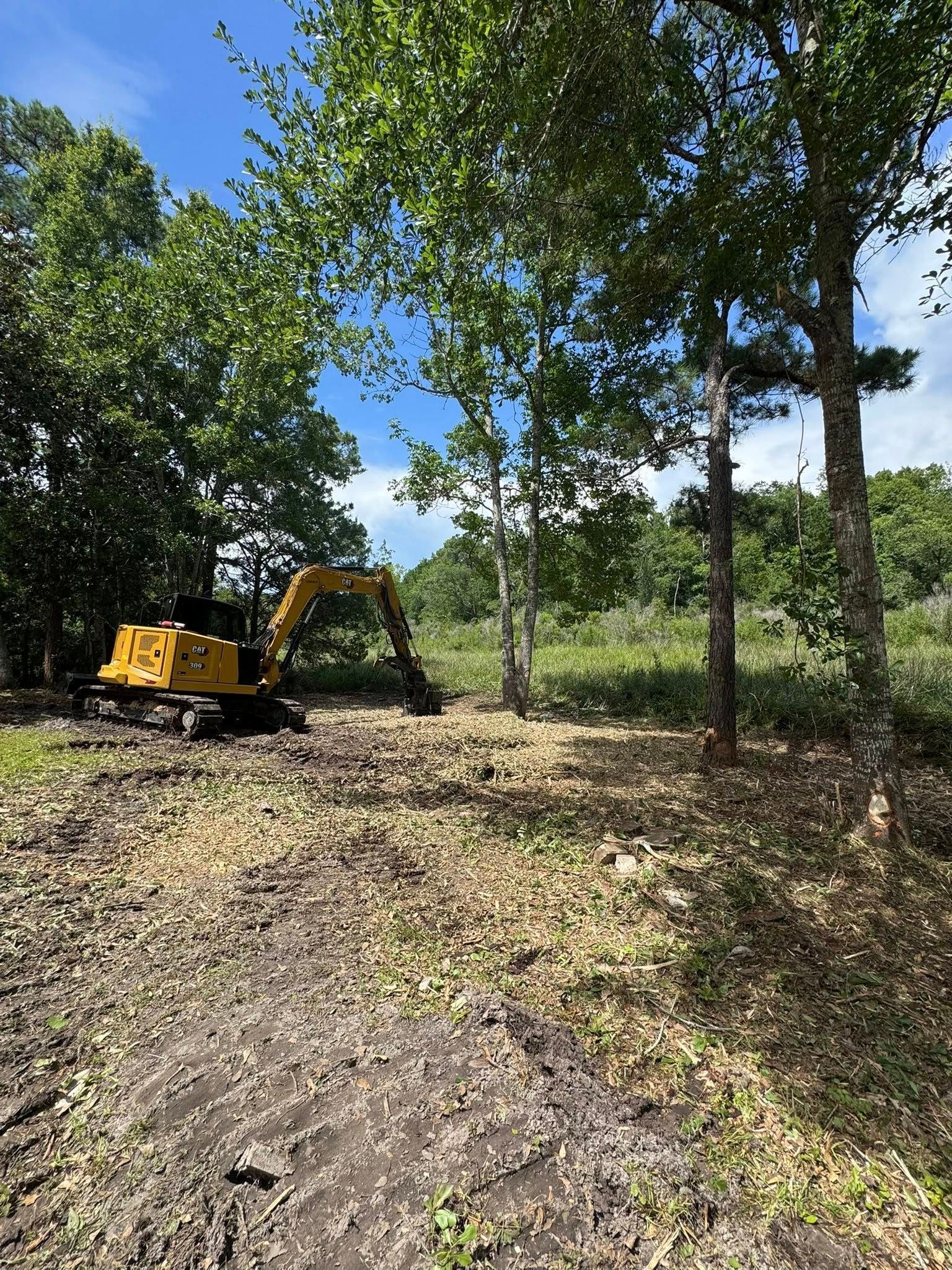 Yellow excavator clearing land near trees under a blue sky.