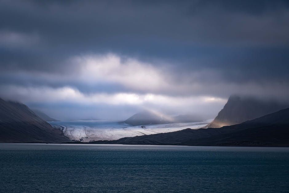 Dark cloudy sky over a glacier and a body of water, mountains in the background.