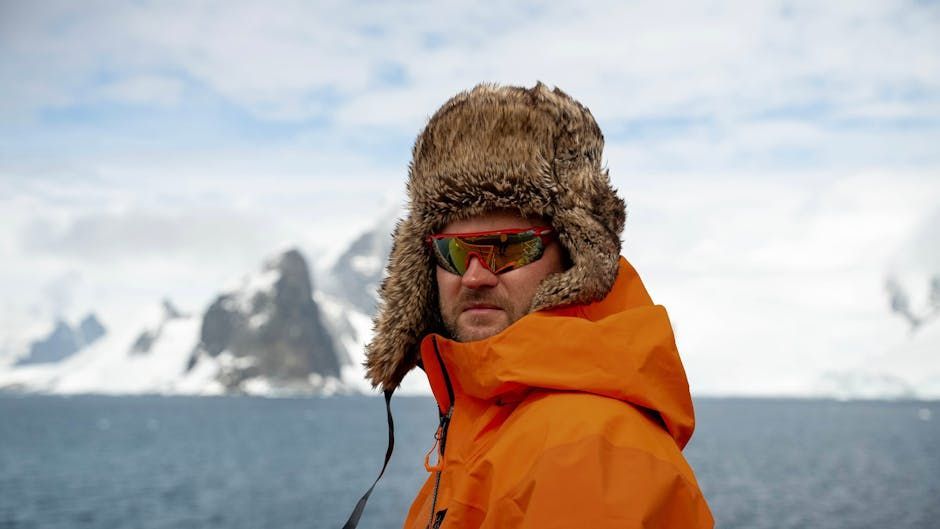 Man in orange parka and fur hat, wearing sunglasses, poses near Antarctic mountains and sea.
