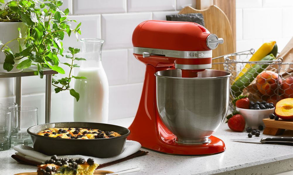 A red kitchenaid mixer is sitting on a counter next to a pan of food.