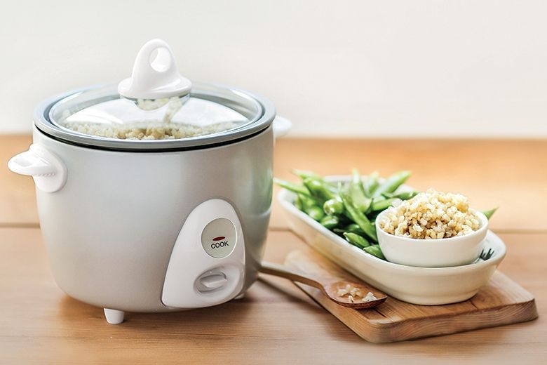 A rice cooker is sitting on a wooden table next to a bowl of rice and green beans.