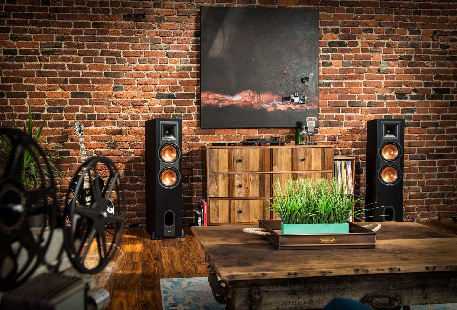 A living room with a brick wall , a wooden table , two speakers and a painting on the wall.