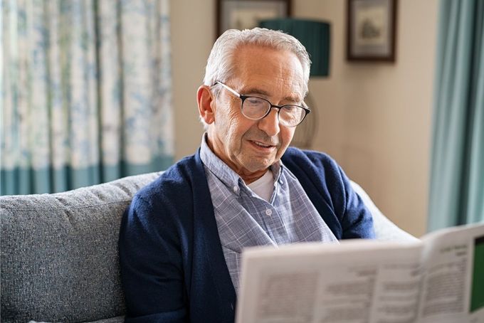 Older man wearing glasses, sitting on a sofa and reading a newspaper.