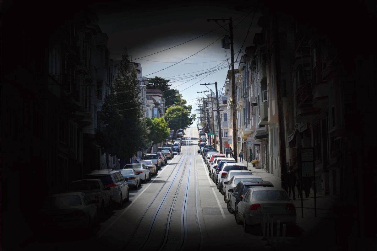 Street lined with parked cars, buildings, and trees. Light flares outward from the center.