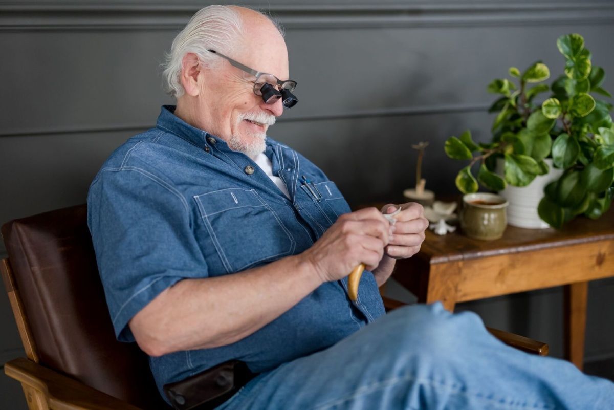 Elderly man in denim shirt and his SVAG eyeglasses smiles while knitting. 
