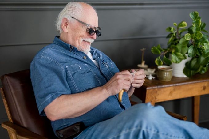 Elderly man in denim shirt and his SVAG eyeglasses smiles while knitting. 